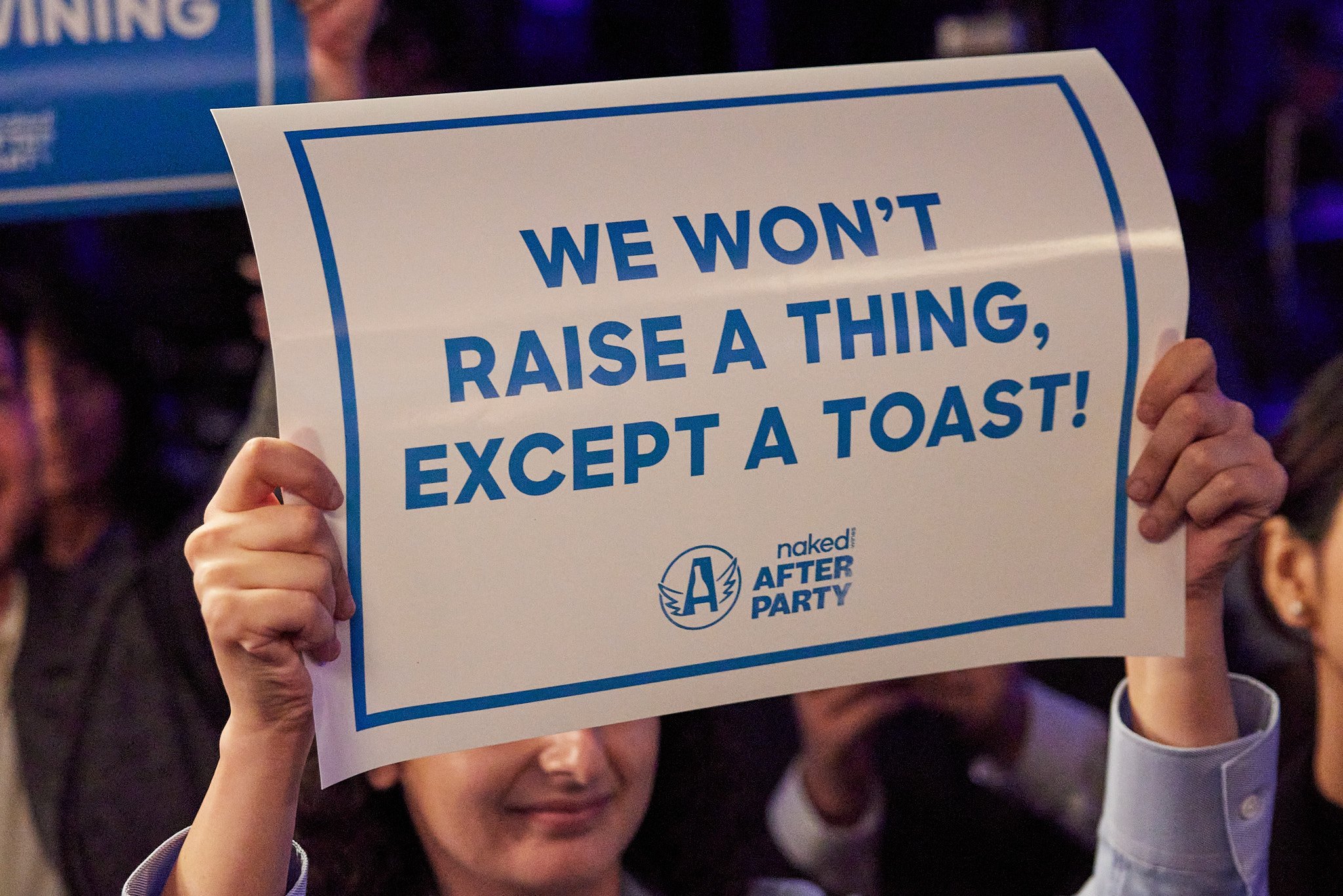 Person holding a sign that says, "We won't raise a thing, except a toast!" at the Naked After Party event.