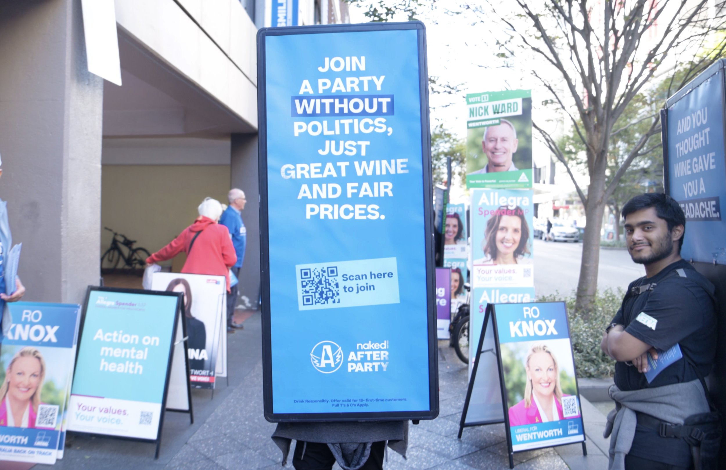 A large blue vertical sign with white text reads, 'Join a party without politics, just great wine and fair prices,' and includes a QR code with a label 'Scan here to join.' Nearby, political campaign posters and signs are displayed, including a smili