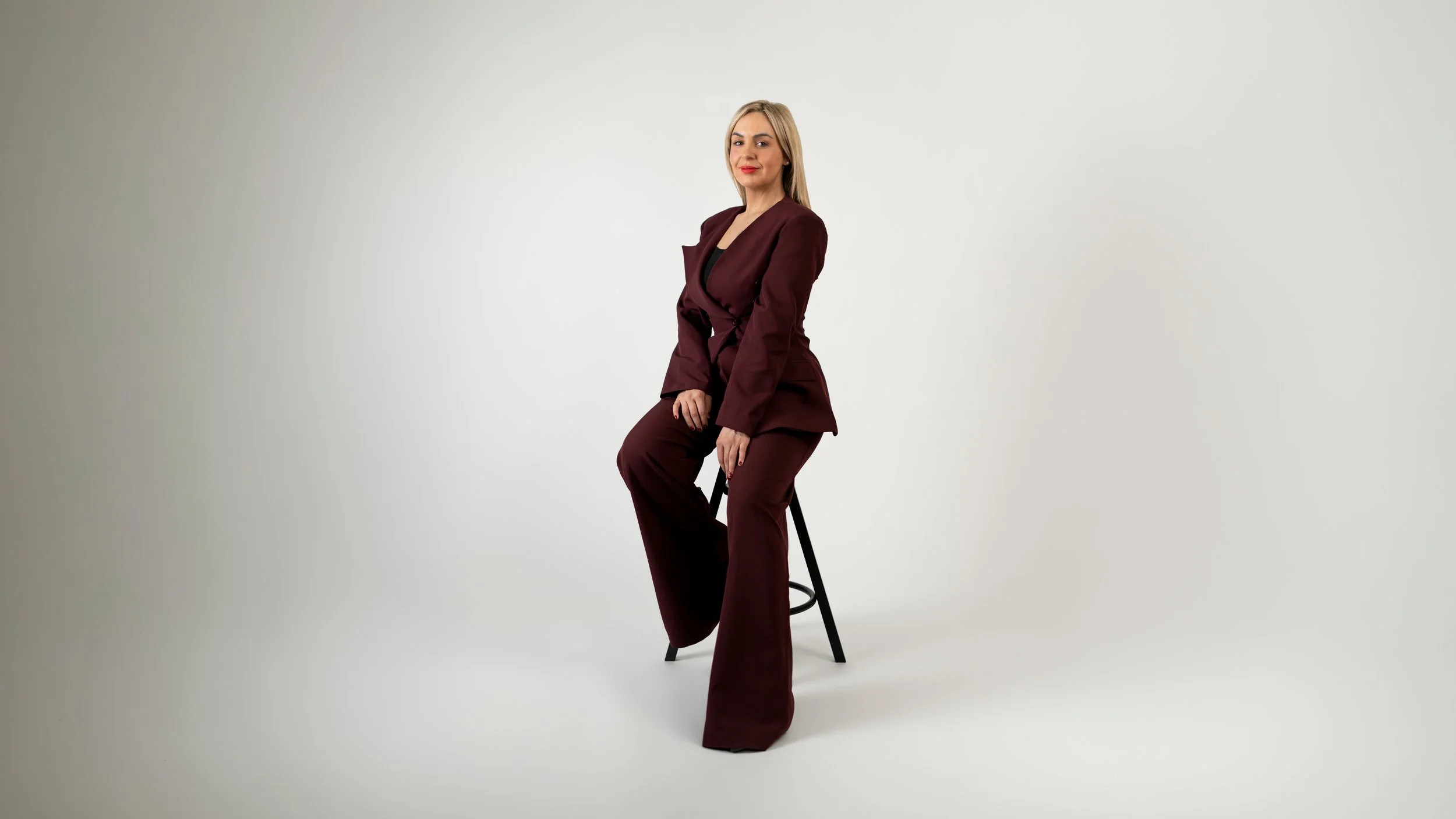 A woman in a dark burgundy business suit sitting on a black bar stool against a plain white background.