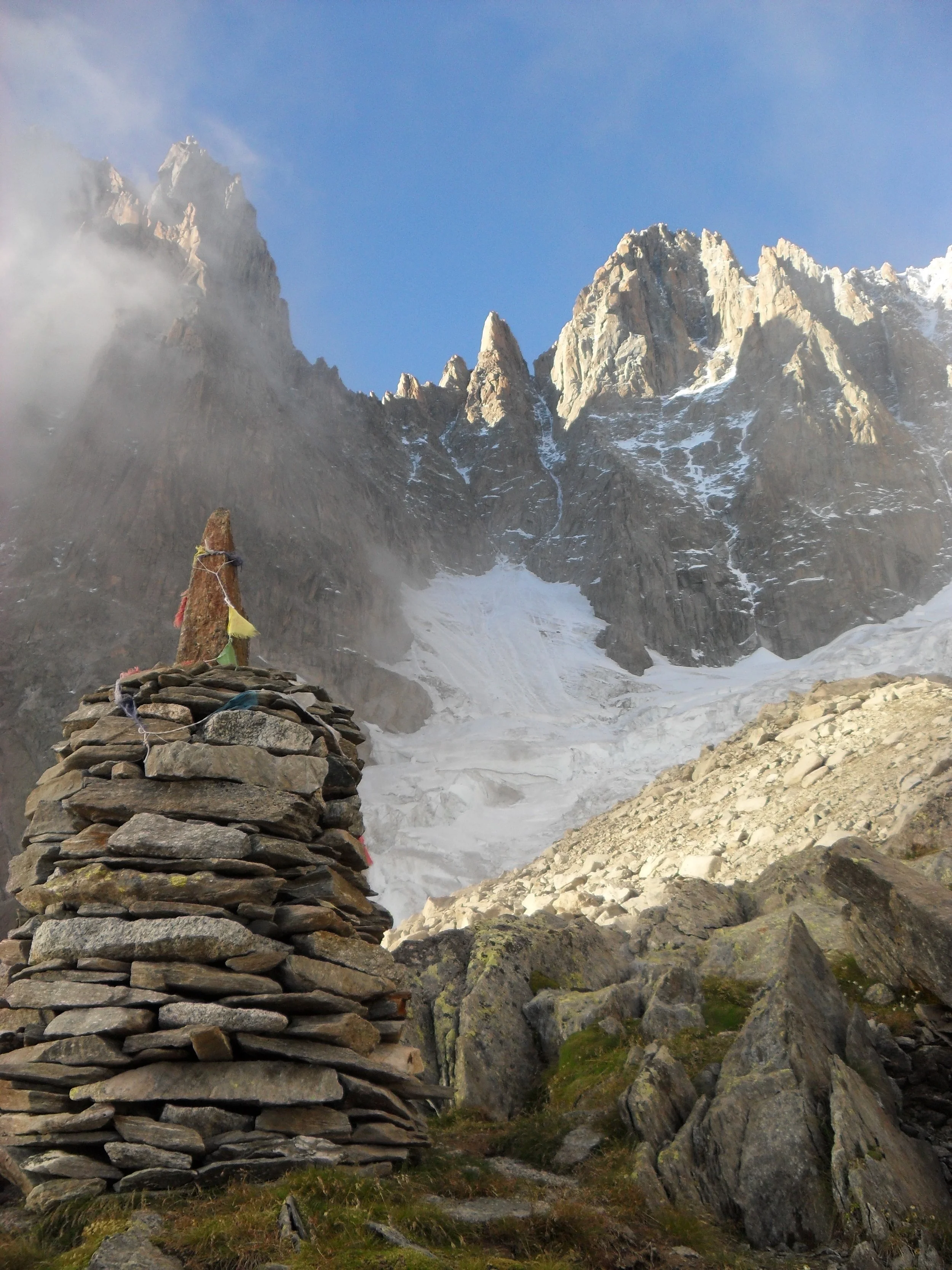 Mountain landscape with rugged peaks, snowy glacier, and a stone cairn with colorful prayer flags