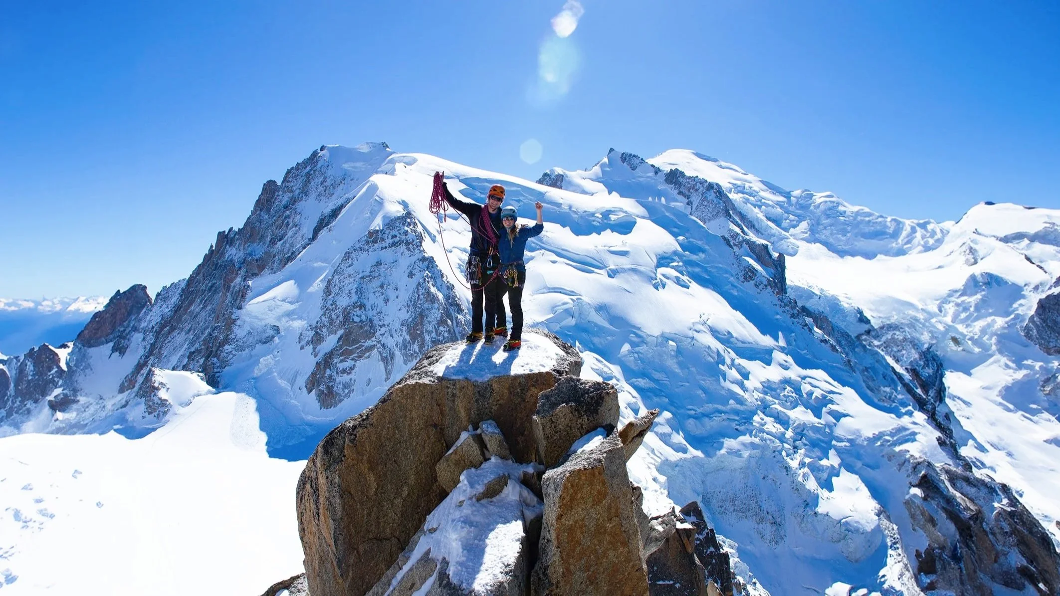 Two climbers standing on a rocky ledge at the summit of a snow-covered mountain, with a backdrop of rugged, snow-capped peaks under a clear blue sky.