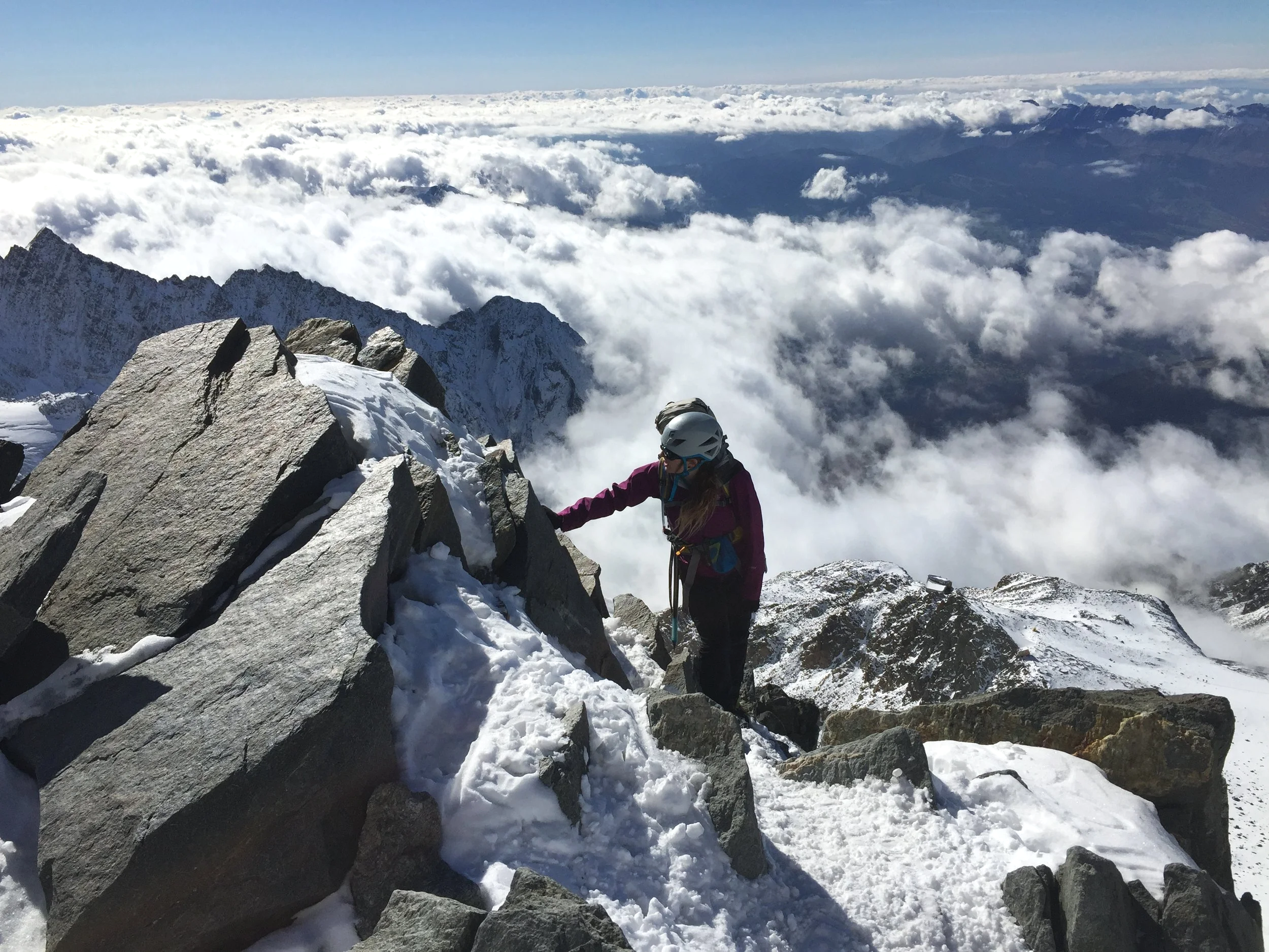 Climber wearing helmet and purple jacket ascending snow and rocks on a mountain ridge above clouds.