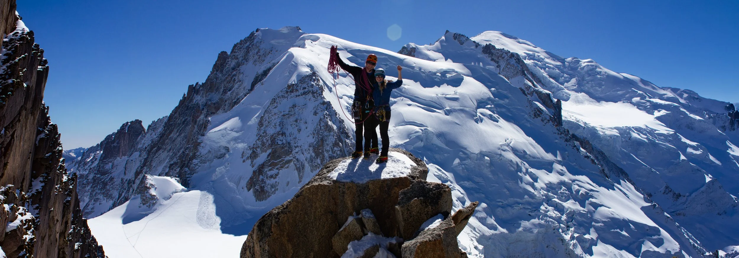 Two climbers standing on a large rock at the top of a snowy mountain, celebrating with raised fists and holding climbing gear, with snow-covered peaks in the background under a clear blue sky.
