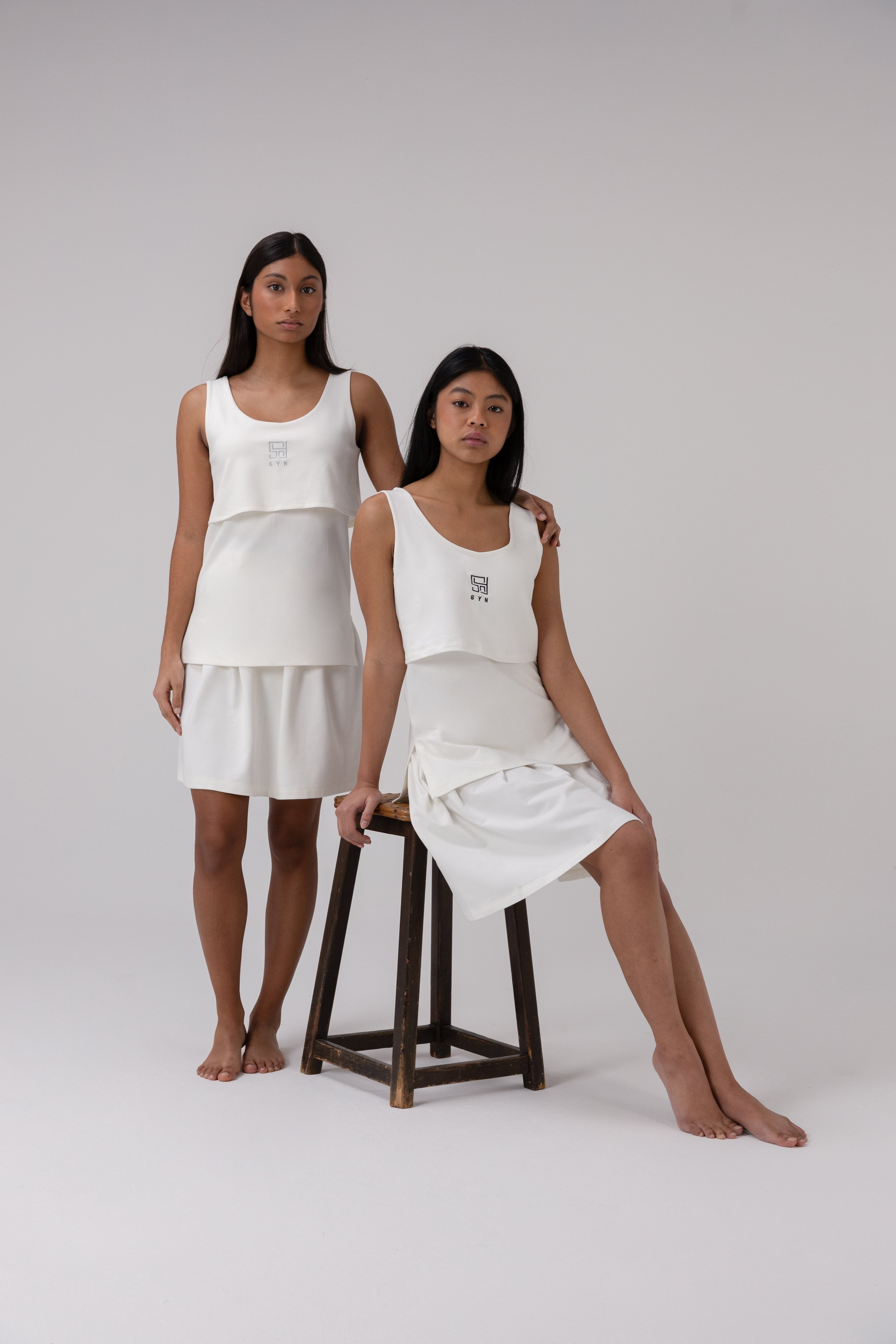 Models wearing SYN STUDIOS white dresses in minimalist studio setup.