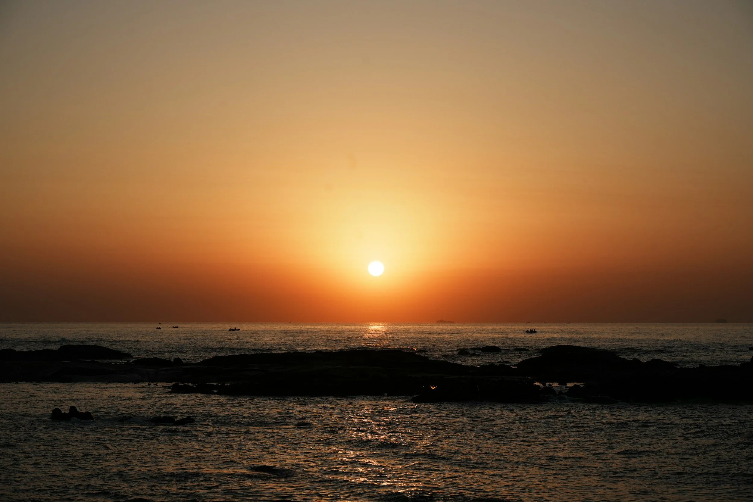 Sunset over the ocean with a few small boats and rocky outcroppings in the foreground.