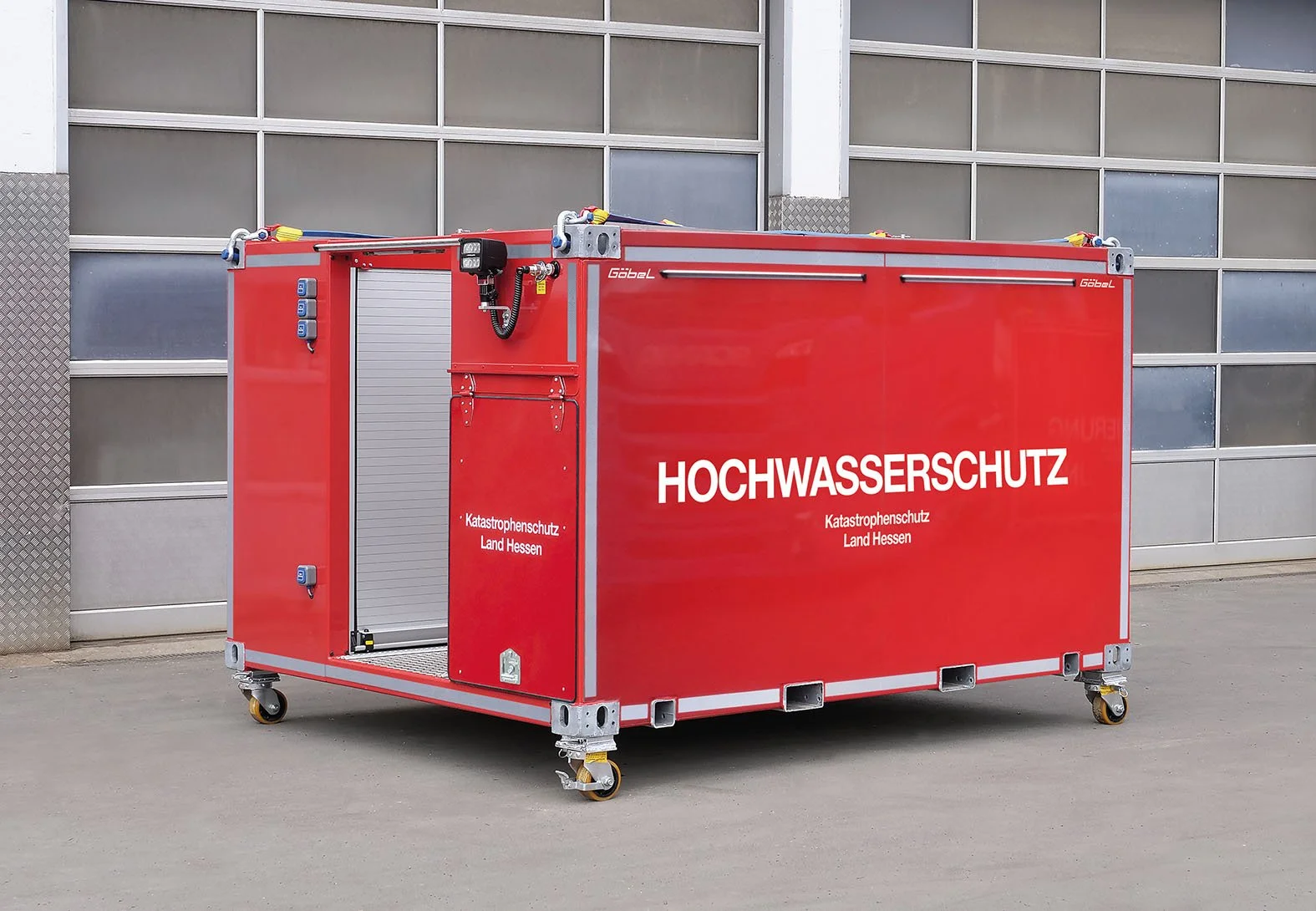 Ein rotes Hochwasserschutz-Container, der für Katastrophenschutz in Hessen verwendet wird, auf Rollen stehend vor einer Gebäude-Fassade.