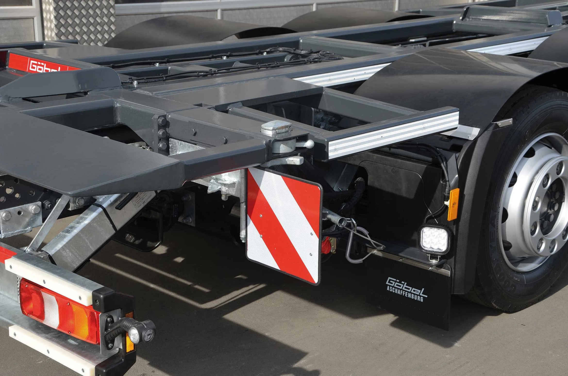 Nahaufnahme eines LKW-Anhängers, der mit einem schwarzen Fahrzeug verbunden ist. Der Anhänger ist grau mit roten und weißen reflektierenden Markierungen, und es sind technische Komponenten sichtbar. Es gibt auch einen Reifen auf der rechten Seite.