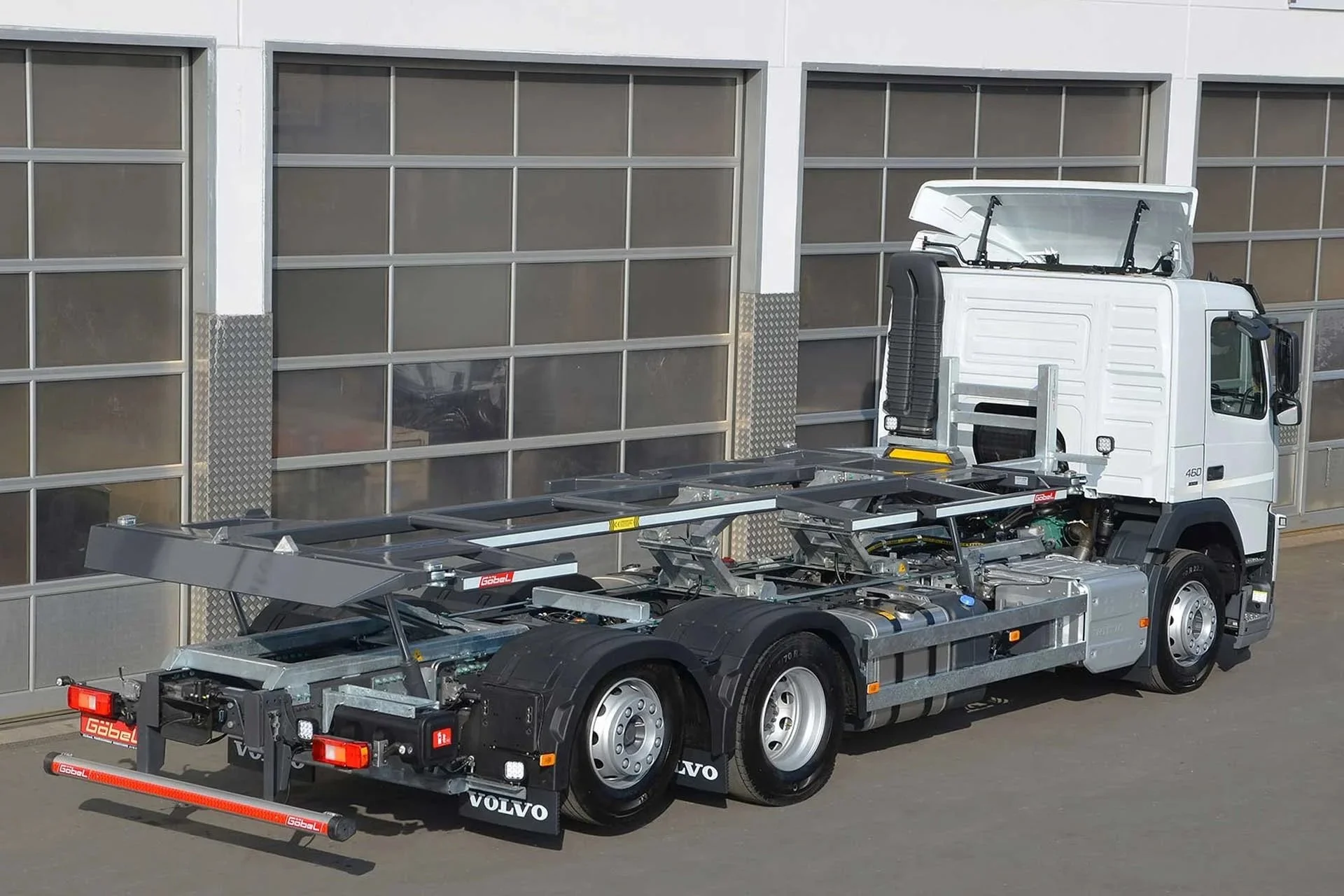 Leeres Auto-Transportfahrzeug mit offener Ladefläche und weißem Lkw-Fahrzeug im Hintergrund, geparkt vor einer modernen Gebäudewand mit großen Fenstern.