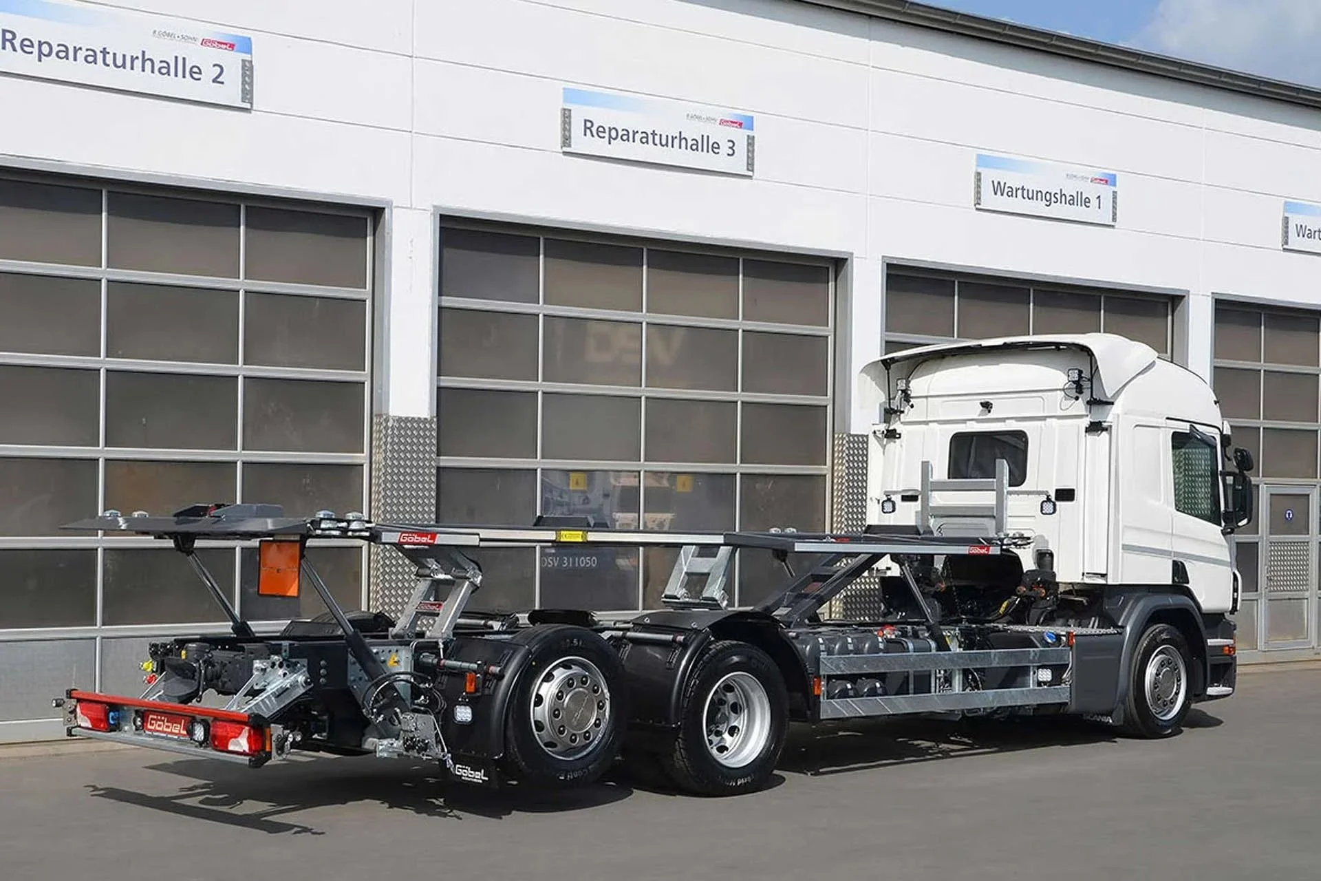 Ein weißer LKW ohne Auflieger, der spezialisierte Fahrzeugtechnik tragt, steht vor einer modernen weißen Werkhalle mit mehreren Beschilderungen. Das Fahrzeug trägt auf dem Rahmen verschiedene Werkzeuge und Geräte.