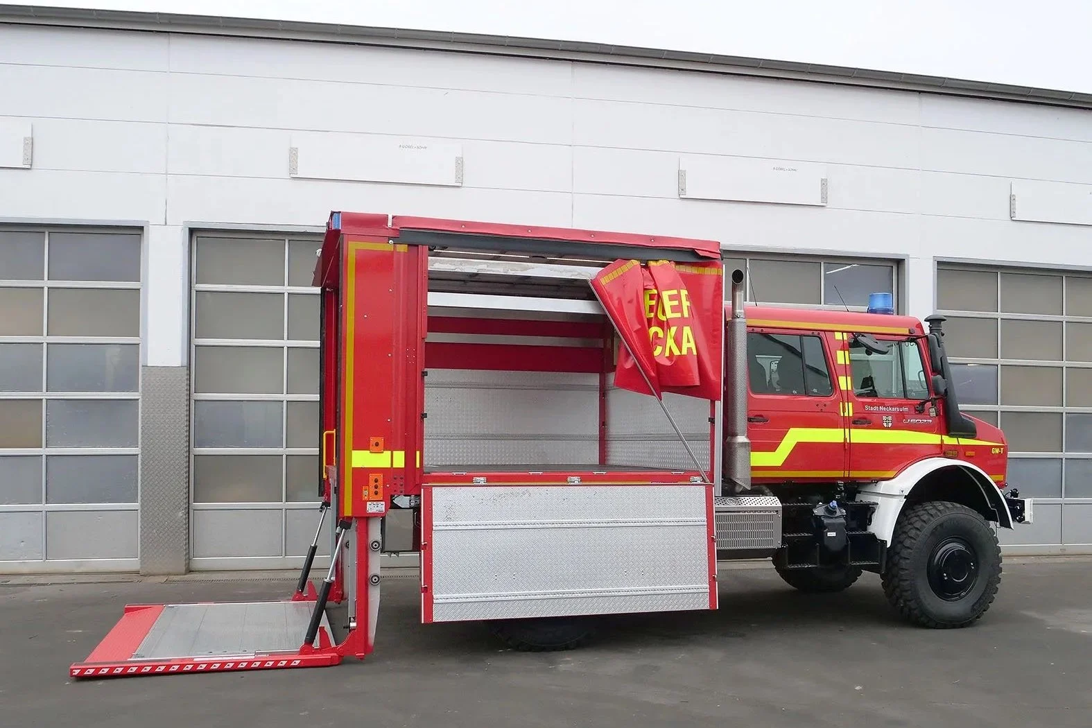Gerätewagen Logistik Mercedes Benz Unimog für Feuerwehr Neckarsulm aufgebaut von Göbel Fahrzeugbau, seitliche Heckansicht mit runtergelassener Ladebordwand und geöffneter rechten Seite