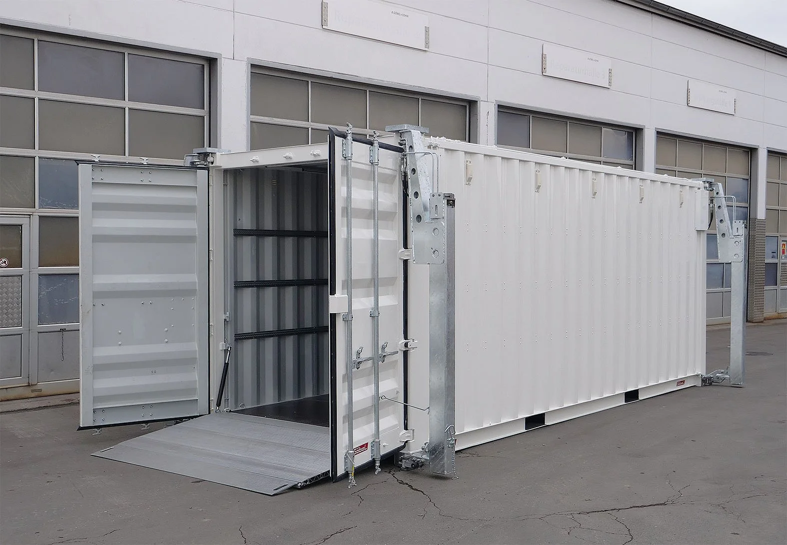Ein mobiler Aufzug mit einer Hubluke, angebracht an einem weißen Container, auf dem Boden vor einer Industriehalle.