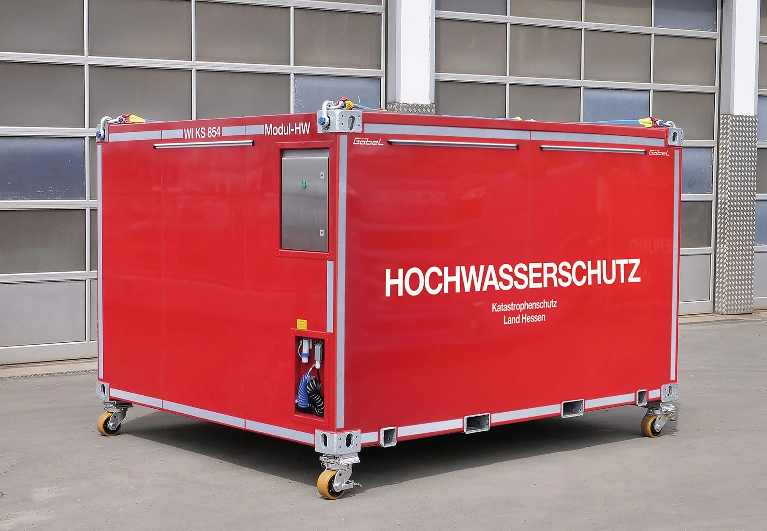 Ein rotes Hochwasserschutzcontainer mit Rollen. Das Container trägt die Beschriftung "HOCHWASSERSCHUTZ" und "Katastrophenschutz Land Hessen". Aufgebaut für das Land Hessen durch Göbel Fahrzeugbau.