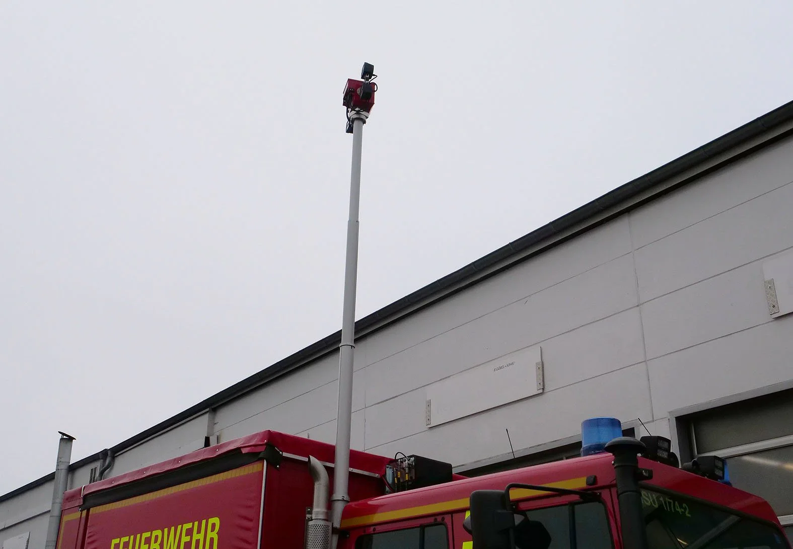 Feuerwehrfahrzeug mit einer aufgestellten Leiter und Warnsirene, vor einem modernen Gebäude mit grauer Fassadenseite.