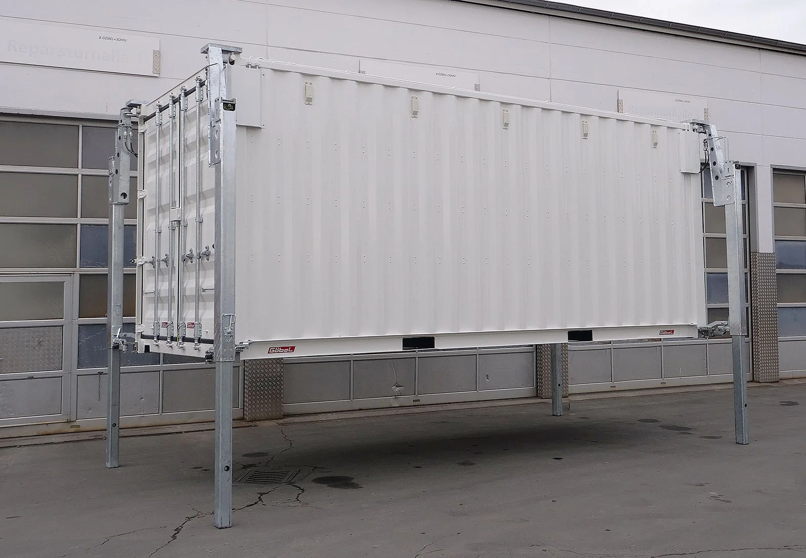Ein großes weißes Container auf einem erhöhten Rahmen außerhalb eines Gebäudes mit einer grauen Wand und großen Fenstern.