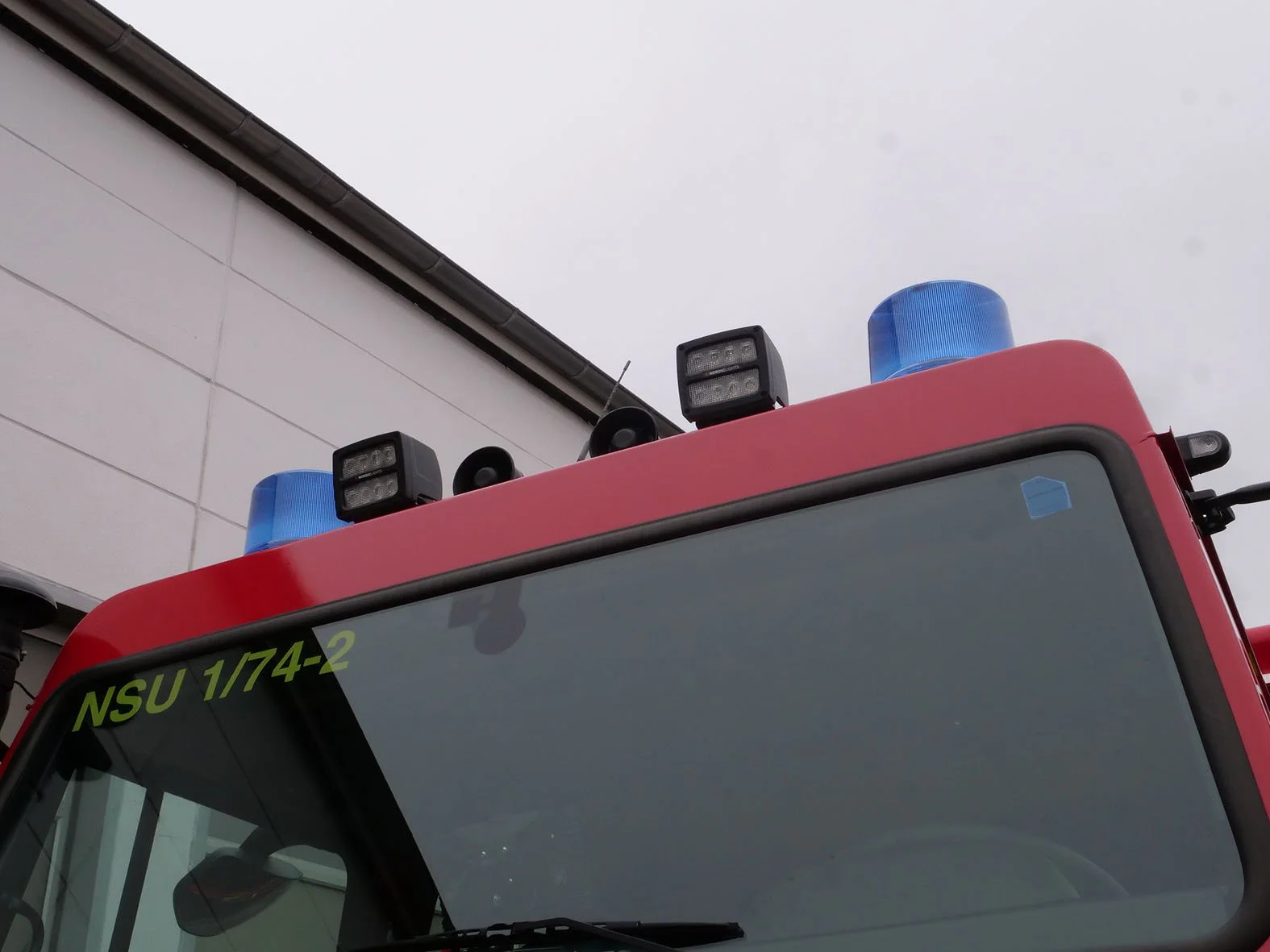 Ein Feuerwehrfahrzeug mit blauen Warnleuchten auf dem Dach, am Himmel ein bewölkter Tag, das Fahrzeug ist vor einem weißen Gebäude geparkt.