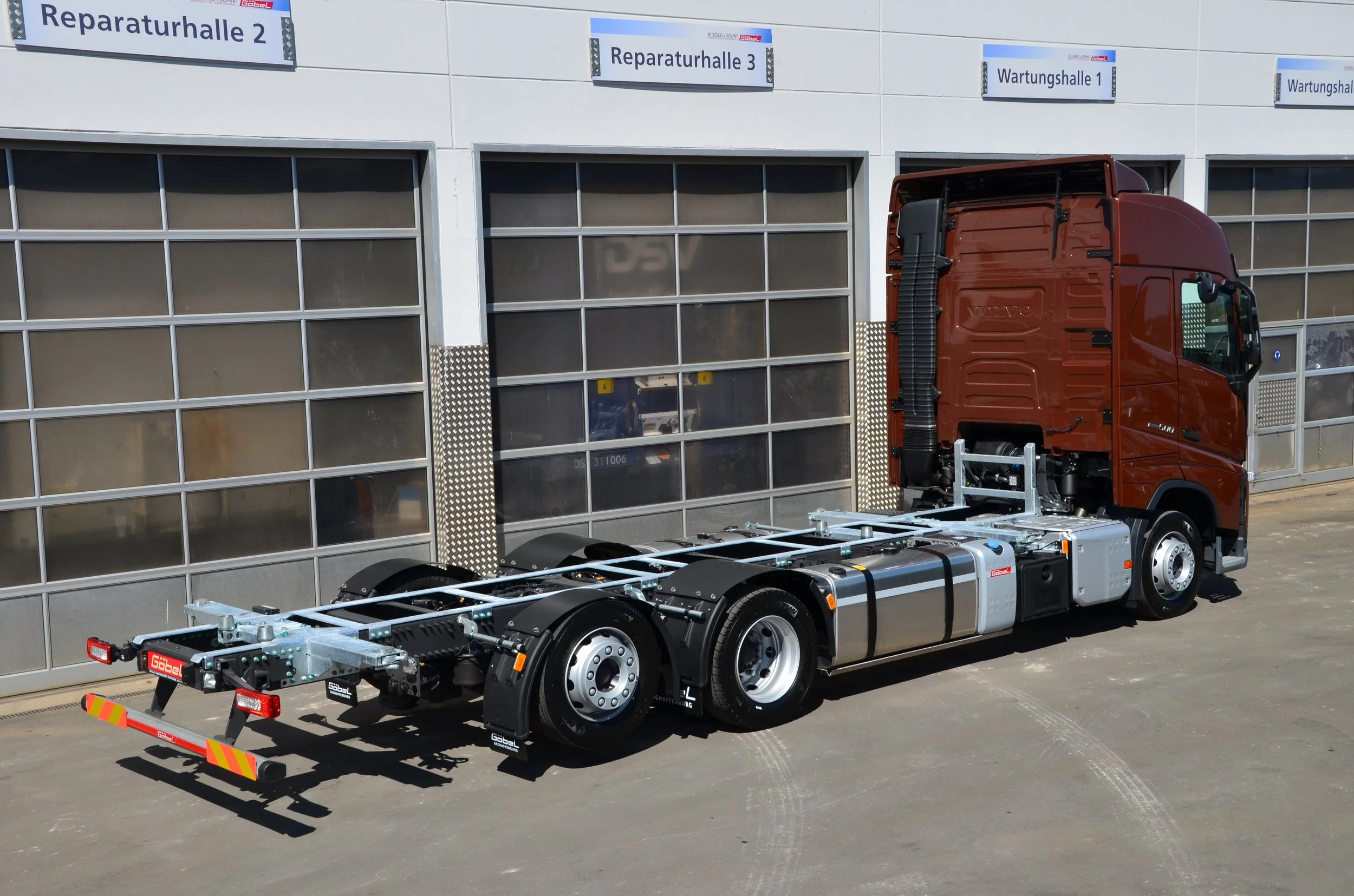 Ein roter LKW ohne Auflieger vor einer modernen Fabrik mit Beschilderungen wie 'Reparaturhalle 2', 'Reparaturhalle 3' und 'Wartungshalle 1'.