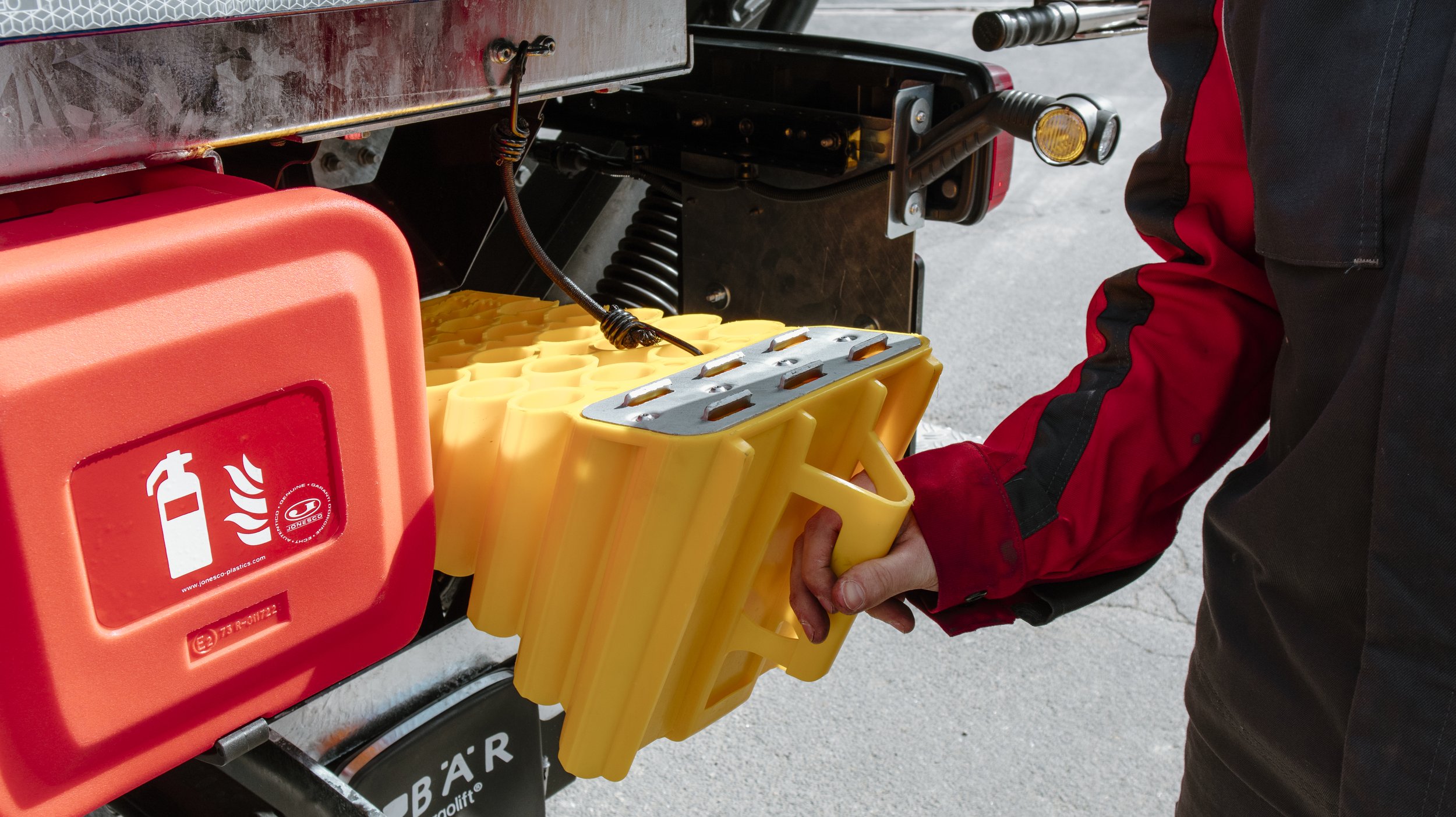 Eine Person in roter und schwarzer Jacke lädt eine gelbe Auffangwanne mit Schutzmittel an einem Feuerlöscher-Container auf einem Fahrzeug. Auf dem roten Container sind Symbole für Feuerlöscher und Brandbekämpfung zu sehen.