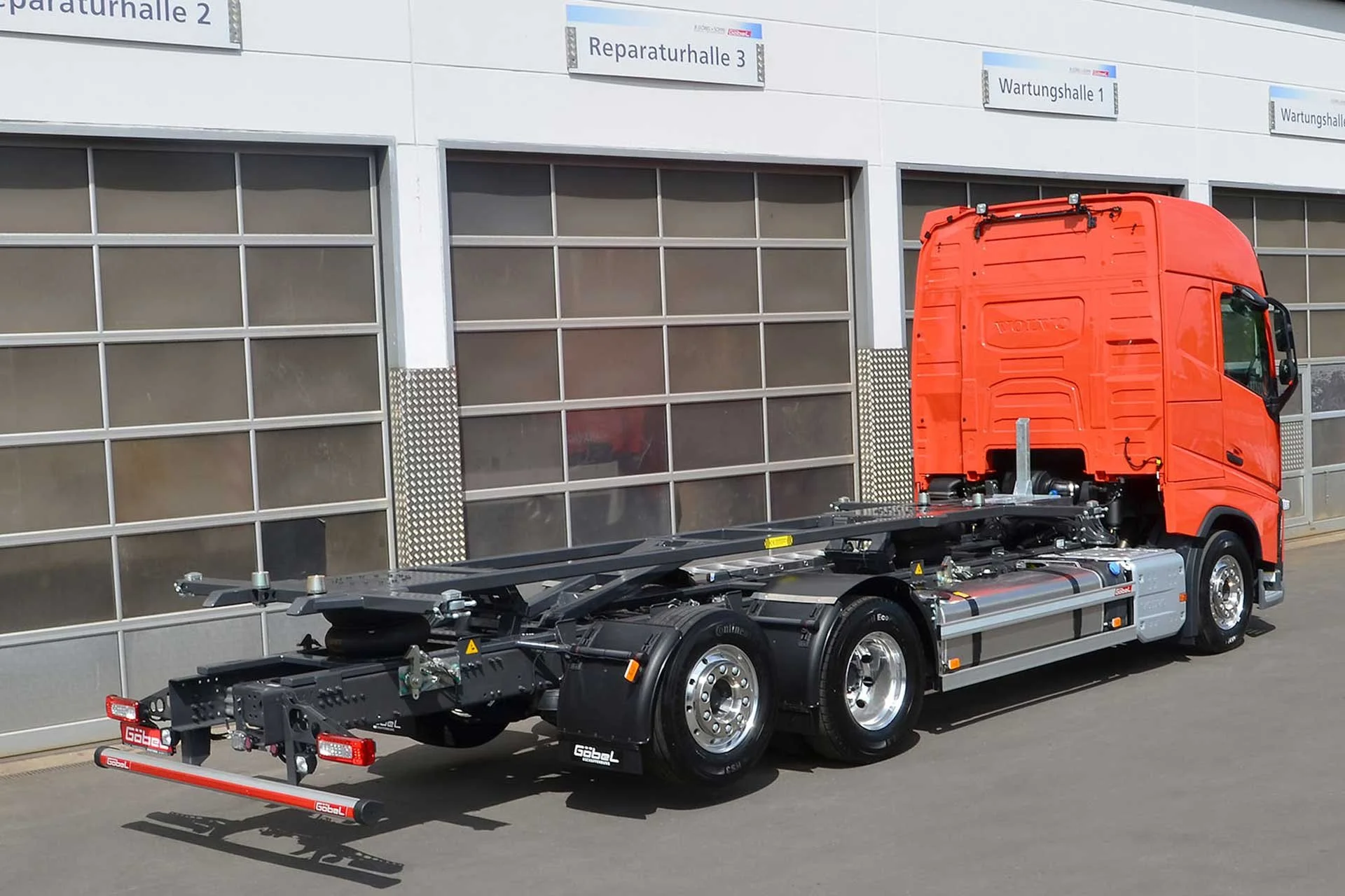 Ein rotes LKW-Chassis ohne Auflieger, geparkt vor einer Gebäude mit beschrifteten Hallen, darunter Werkshalle 1, Reparturhalle 3, Wartungshalle 1.
