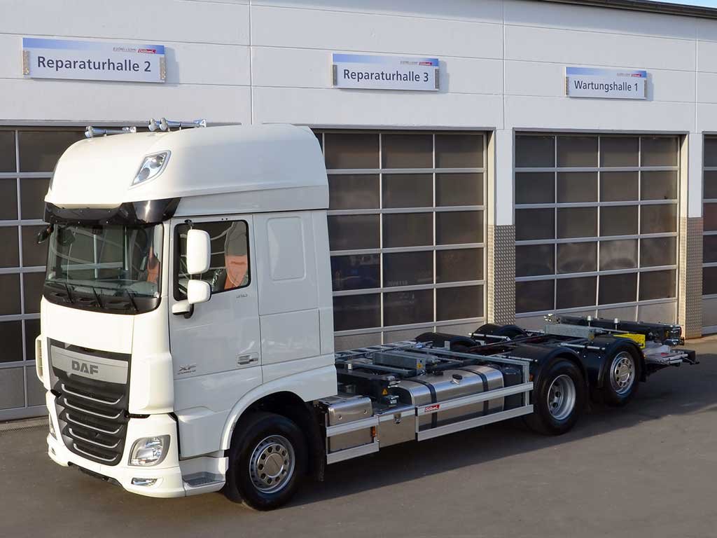 Ein weißer LKW ohne Auflieger, parkt vor einer Werkstatt mit Beschilderung 'Reparaturhalle 2, 3' und 'Wartungshalle 1'.