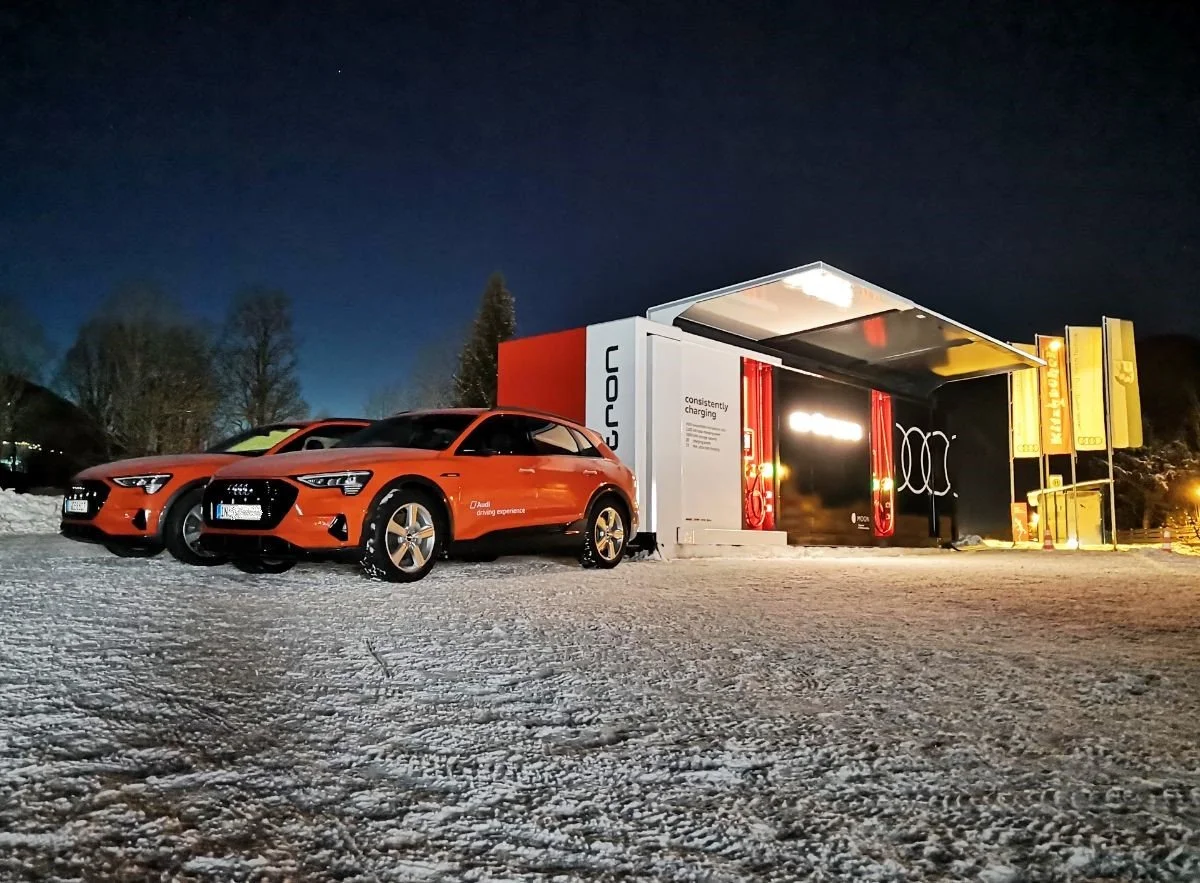 Zwei rote Autos vor einem Ausstellungsstand mit Audi-Logo in einer verschneiten Umgebung bei Nacht.