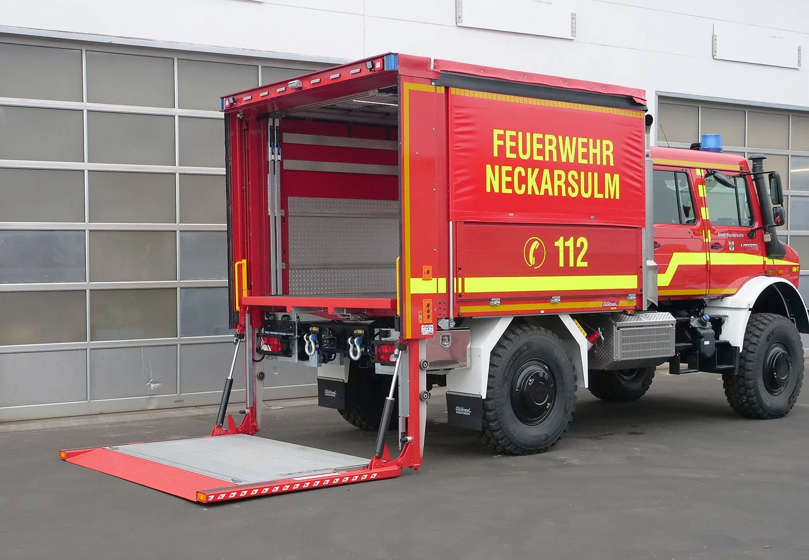Feuerwehrfahrzeug der Feuerwehr Neckarsulm mit offener Ladefläche und eingebauter Rampe, rot mit gelber Beschriftung und Blaulicht auf dem Dach, parkiert vor einer modernen Gebäude Fassaden