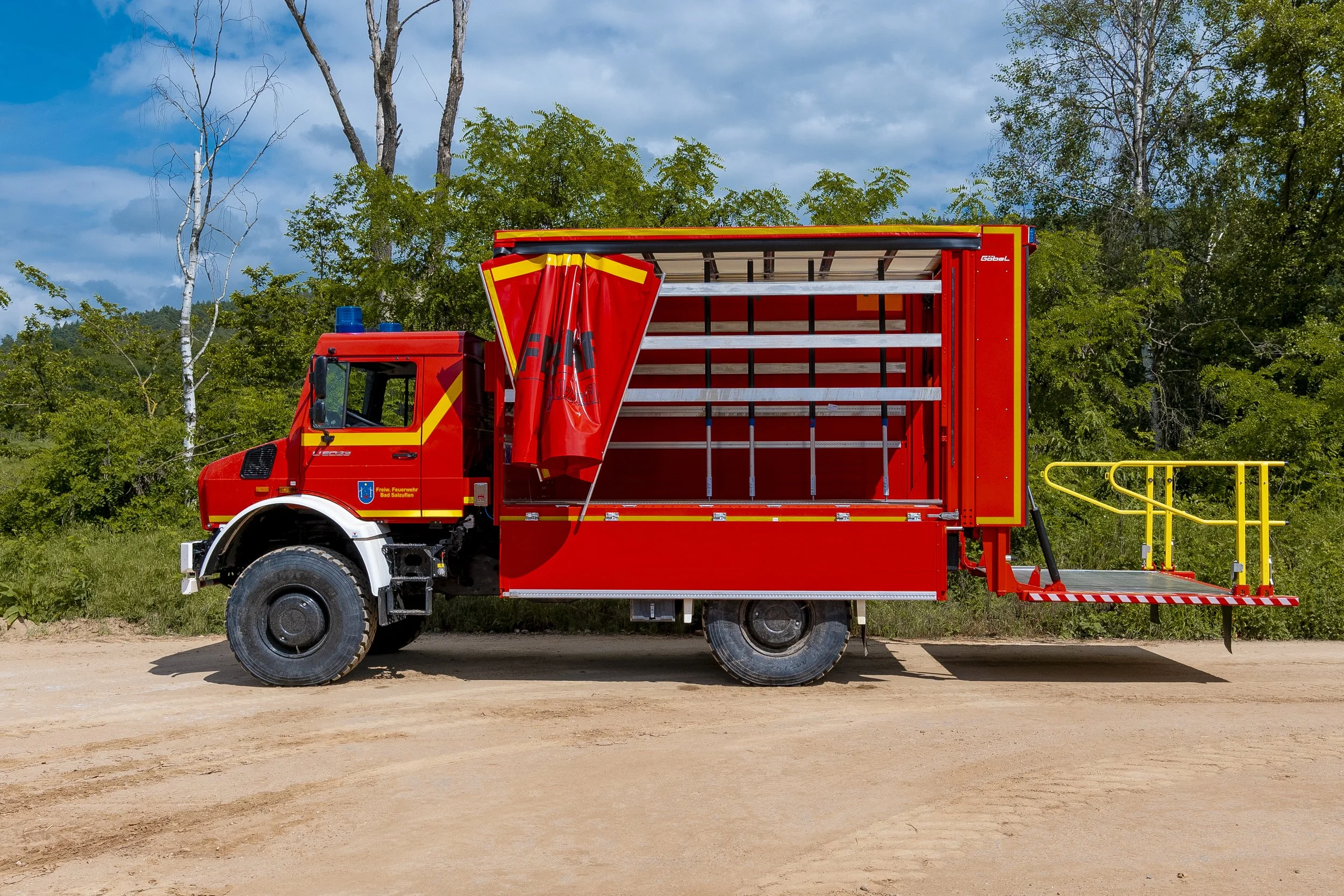 Rotes Feuerwehrfahrzeug mit offenen Laderampe und gelbem Sicherheitsgeländer. Aufbau auf einen Unimog von Mercedes Benz durch die Firma Göbel Fahrzeugbau.
