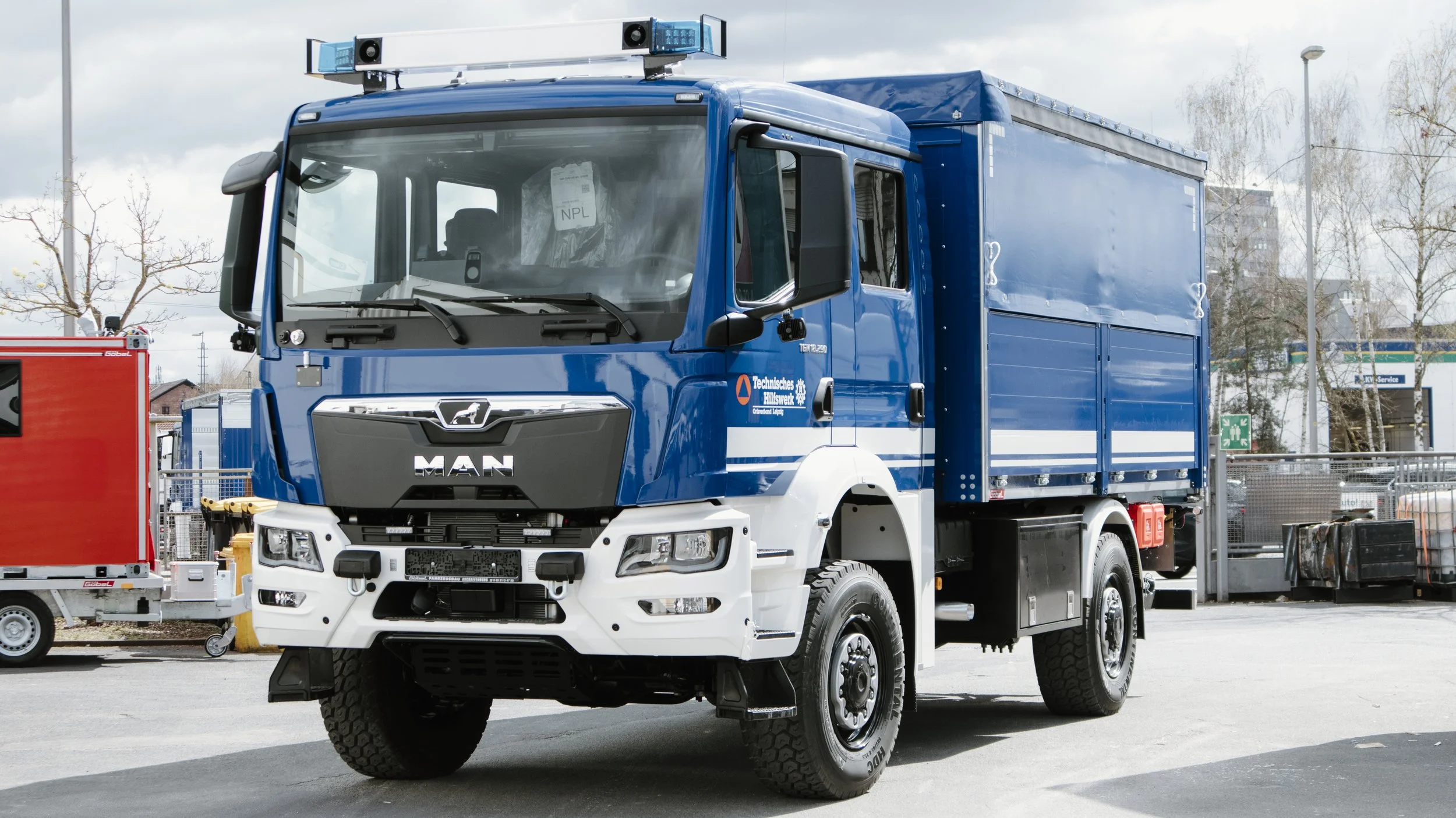 Ein großer blauer MAN-LKW mit einem Container auf der Ladefläche, der mit einem blauen Planen bedeckt ist, steht auf einem Parkplatz. Es gibt ein kleines rotes Fahrzeug im Vordergrund und einen Zaun sowie Bäume im Hintergrund.