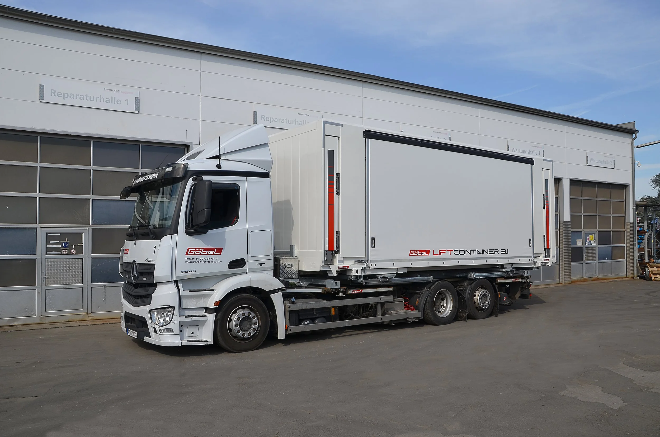 Ein weißer Lkw steht vor einer Fabrikhalle mit mehreren Schildern. Der Lkw hat einen großen Kastenaufbau und ist bereit für den Transport.