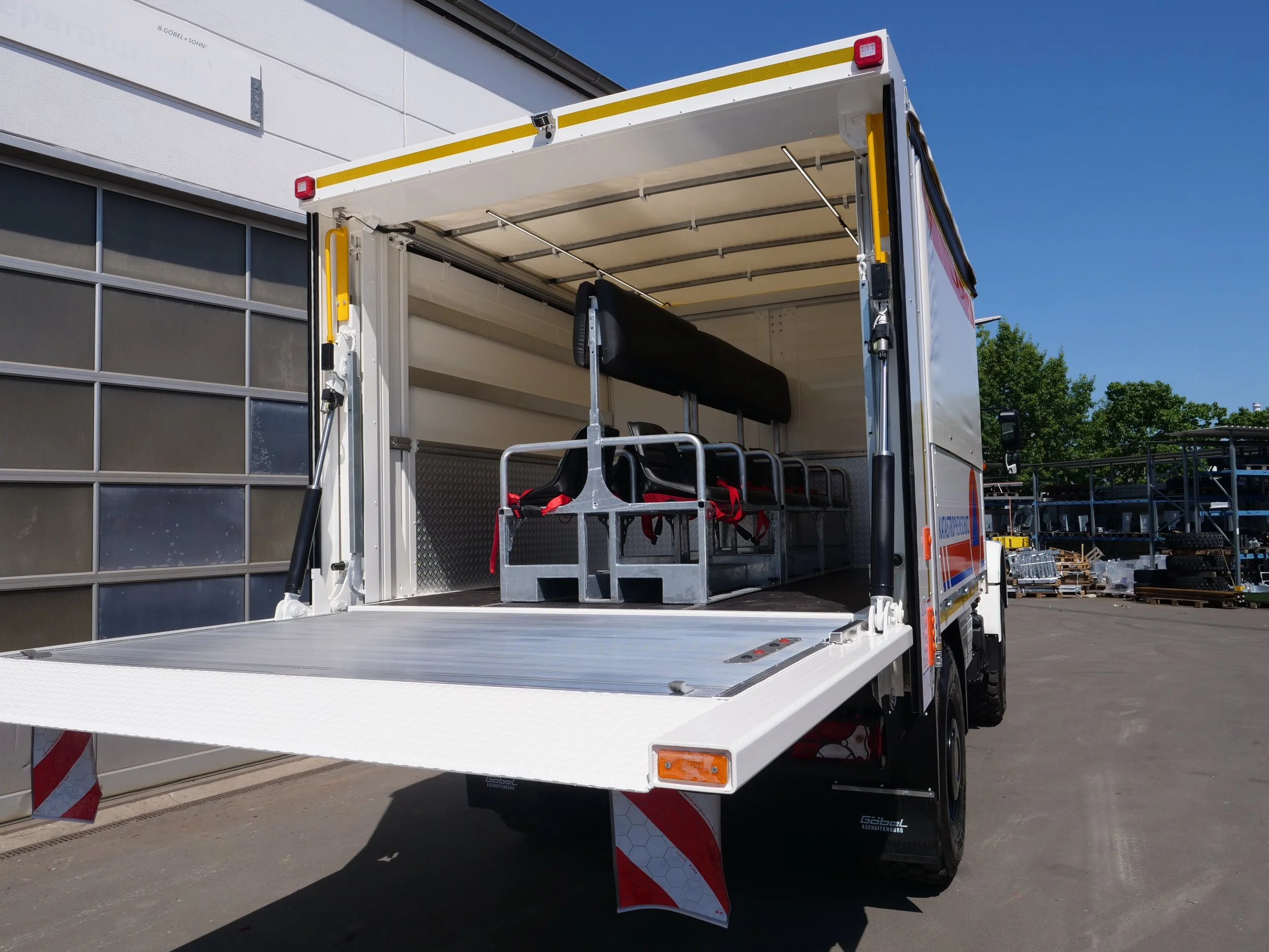 LKW mit offener Ladefläche, die für den Transport vorbereitet ist, steht vor einem modernen Gebäude, blauer Himmel und Bäume im Hintergrund.
