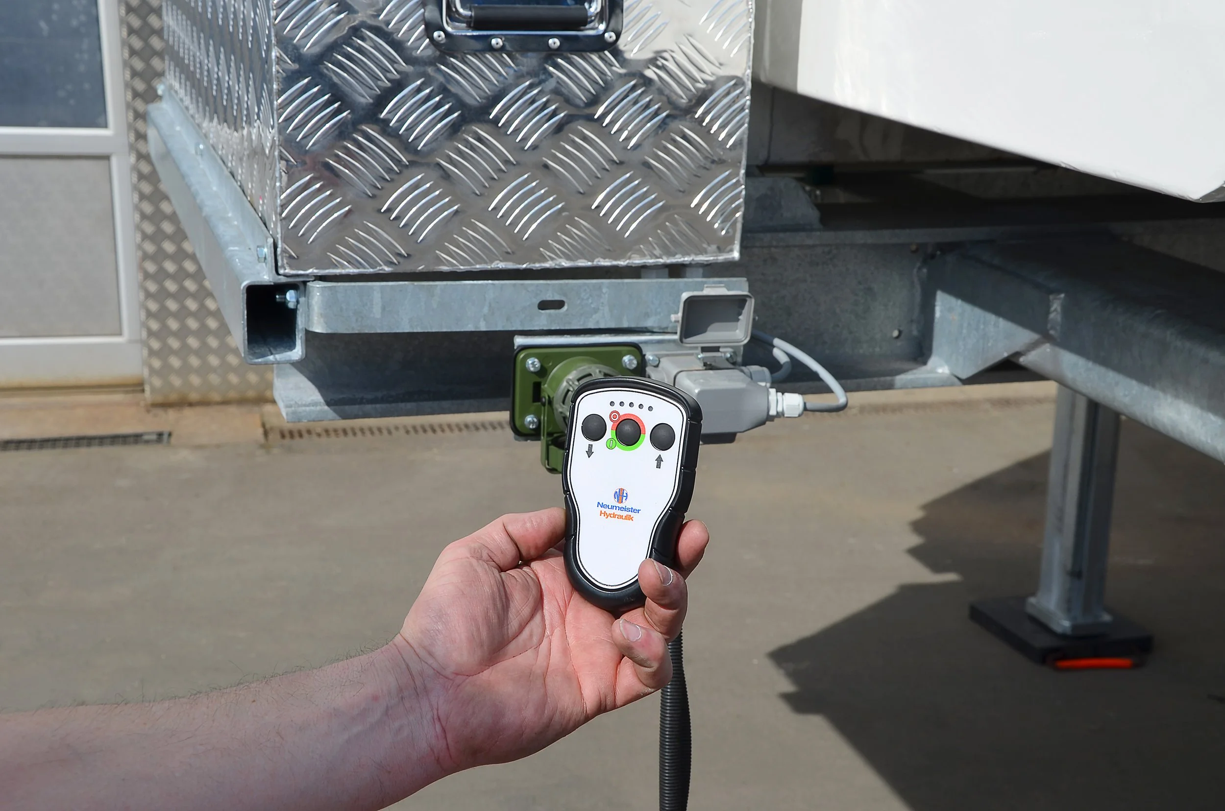 Hand hält eine Fernbedienung vor einem Metallkäfig mit einem elektrischen Gerät, wahrscheinlich eine Hydraulikkonsole, in einem technischen Umfeld.