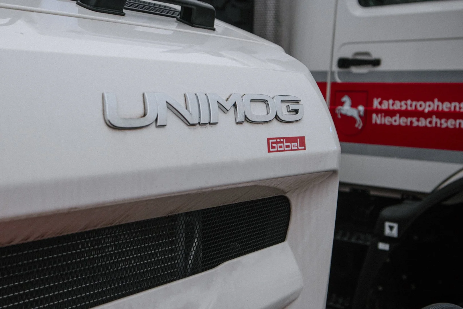 Nahaufnahme eines weißen UNIMOG Fahrzeugs mit einem GOTEL Aufkleber. Im Hintergrund ist ein Fahrzeug mit einem roten Aufkleber mit Weißer Pferde-Logo und der Aufschrift 'Katastrophenschutz Niedersachsen' zu sehen.