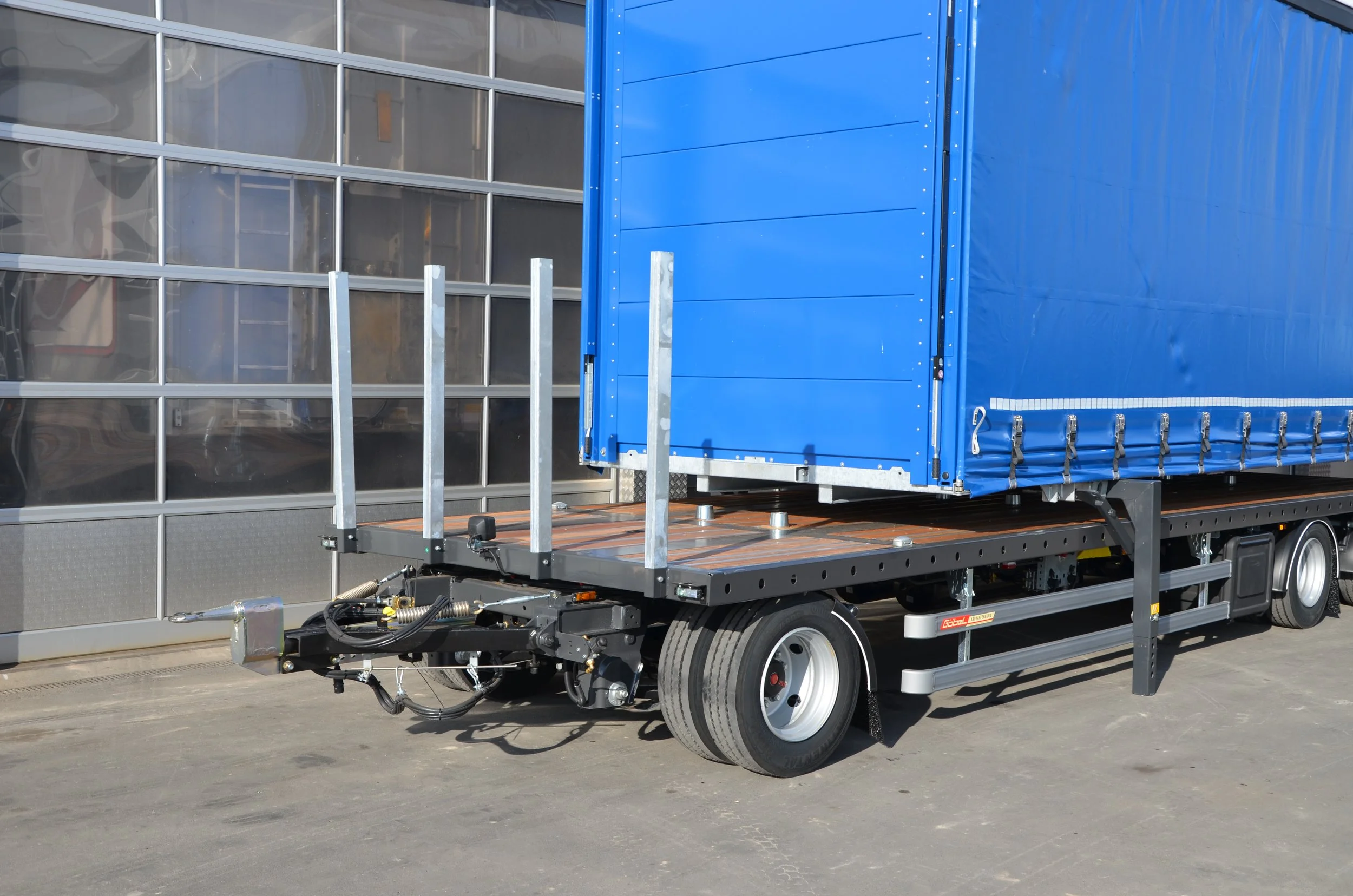 Ein LKW mit blauer Plane auf der Ladefläche ist vor einem Glasgebäude abgestellt. Es sind einige Metallstützen auf der Ladefläche. Der LKW scheint für den Transport von Möbeln oder sperrigem Gütern vorbereitet zu sein.