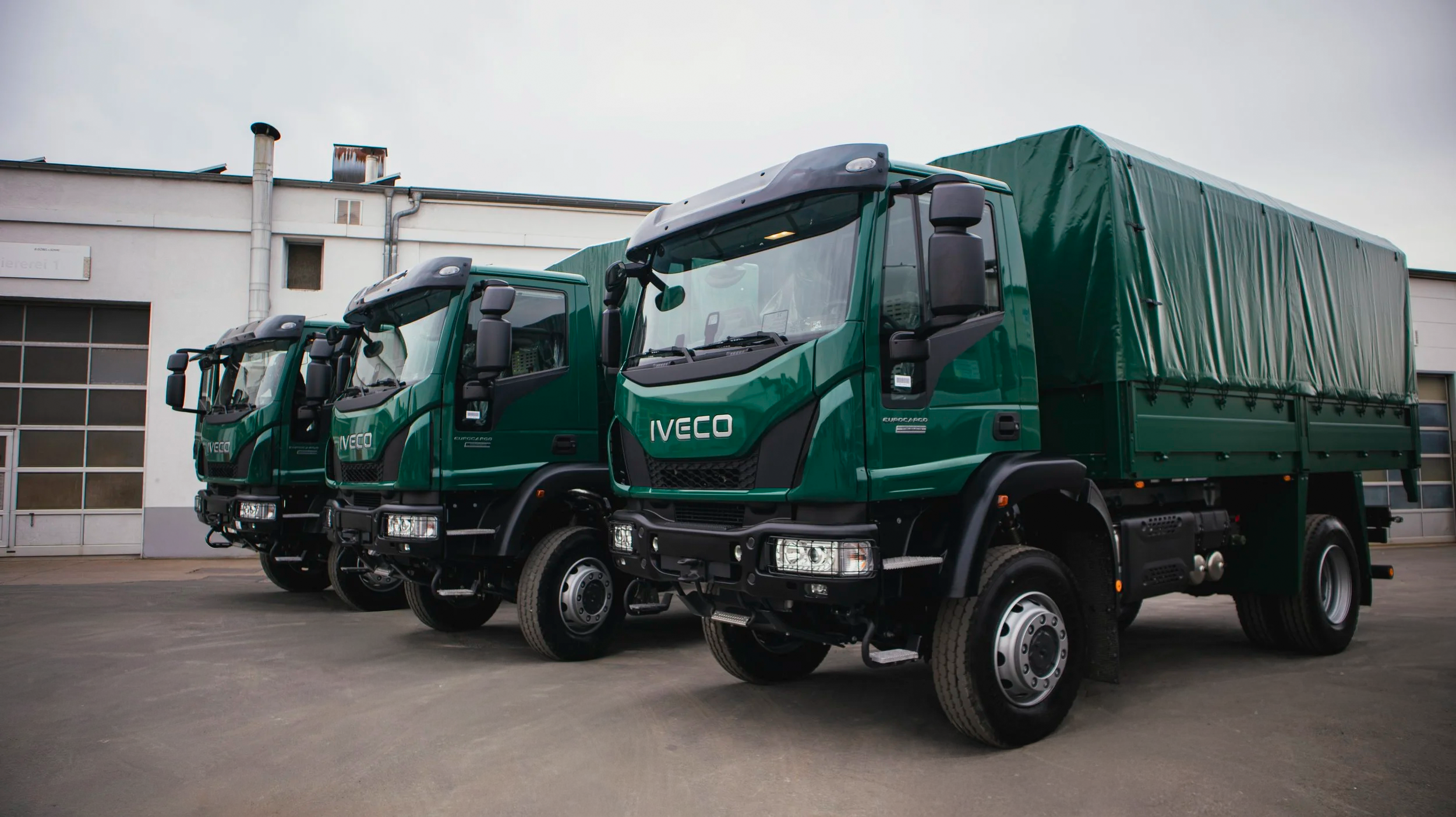 Drei grüne Lastwagen mit dem Markenlogo IVECO stehen nebeneinander auf einem Parkplatz vor einem Gebäude.