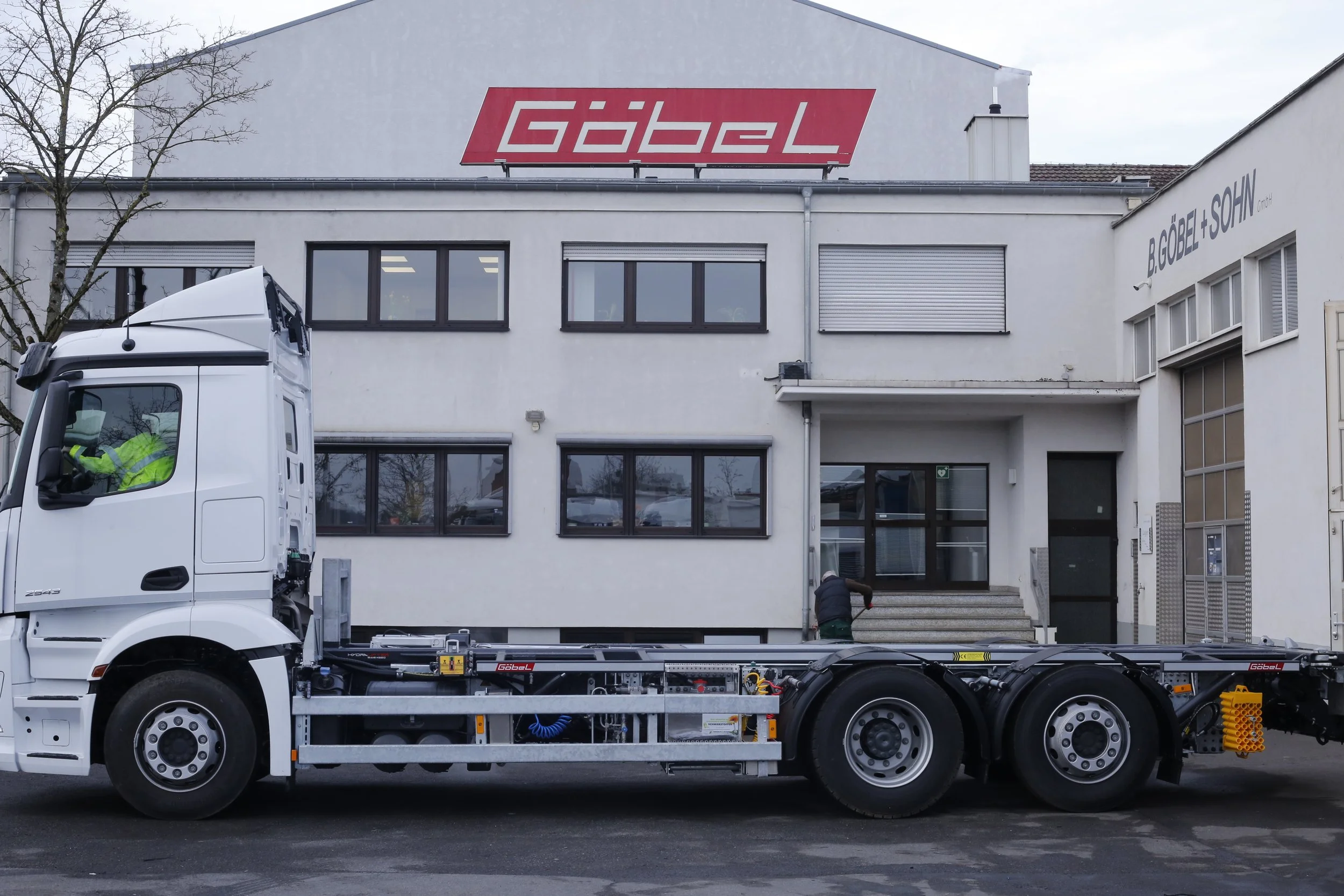 Ein weißer Lastwagen steht vor einem weißen Gebäude mit einem großen roten Firmenlogo 'Göbel'. Es scheint, dass Arbeiten am Fahrzeug durchgeführt werden, möglicherweise Wartung oder Reparatur. Zwei Arbeiter sind sichtbar, einer ist in der Nähe des Gebäudes, der andere arbeitet an dem Fahrzeug. Das Gebäude hat Fenster und einen Eingangstürenbereich auf der rechten Seite.
