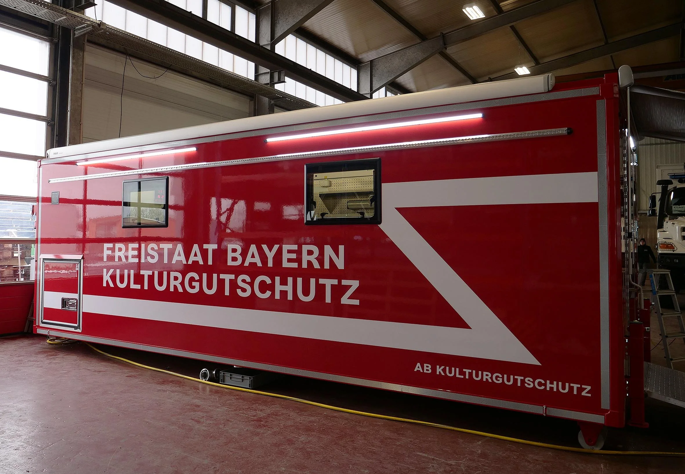 Roter KulturGutshaus Wagen mit weißen und roten Streifen, auf dem steht 'Freistaat Bayern Kulturgutschutz', im Inneren einer Industriehalle.