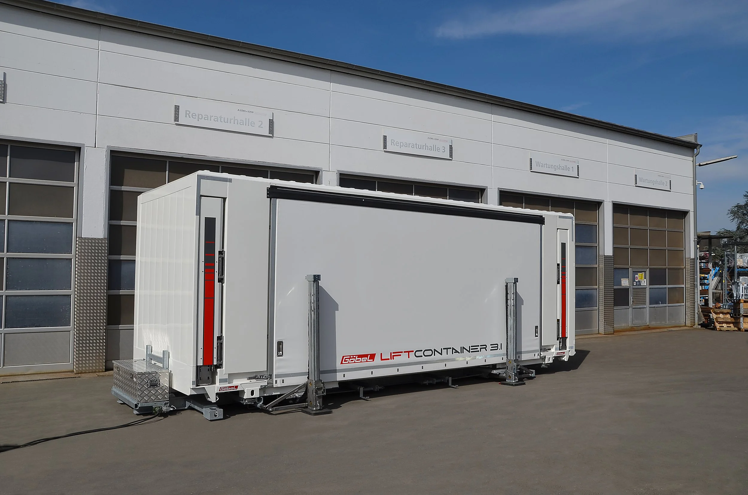 Ein weißer Kofferraumcontainer mit dem Schriftzug G3 Gabel LIFT CONTAINER 3.1 steht vor einer Industriehalle mit mehreren Beschilderungen. Der Himmel ist blau mit wenigen Wolken.