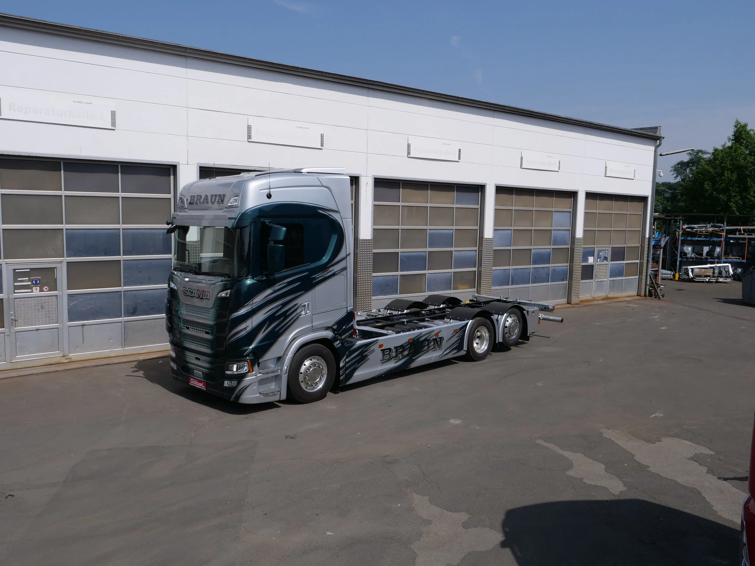 Ein grauer LKW mit der Aufschrift "BRAUN" auf der Seite, parkiert vor einer weißen Garage mit mehreren Toren und einigen Schildern darüber, im Außenbereich an einem sonnigen Tag.