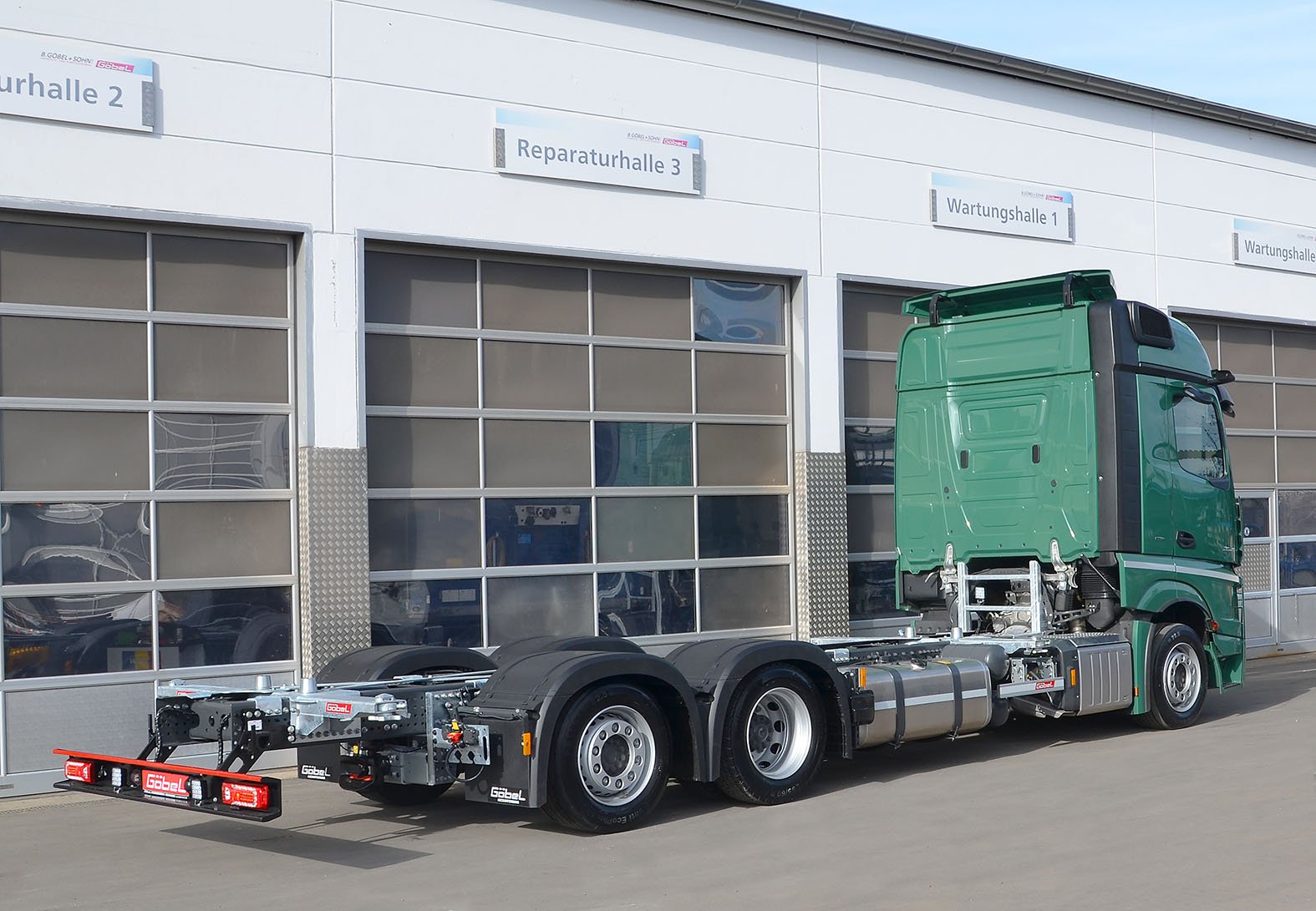 Ein grüner LKW ohne Auflieger vor einer Werkstatt mit Schildern wie 'Reparaturhalle 3' und 'Wartungshalle 1'.