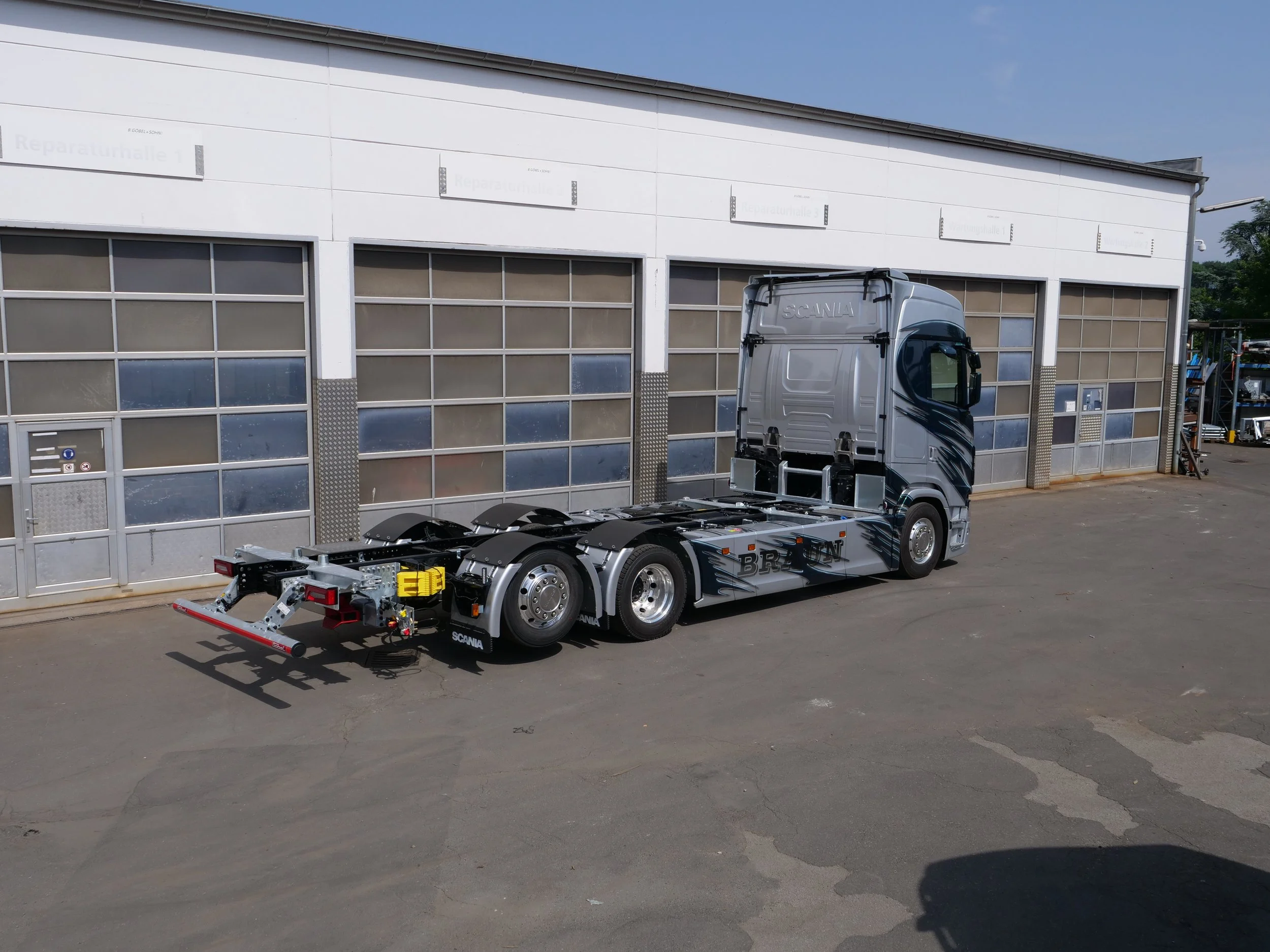 Ein leerer LKW ohne Auflieger vor einer weißen Gebäudefassade mit mehreren Schildern und großen Toren.