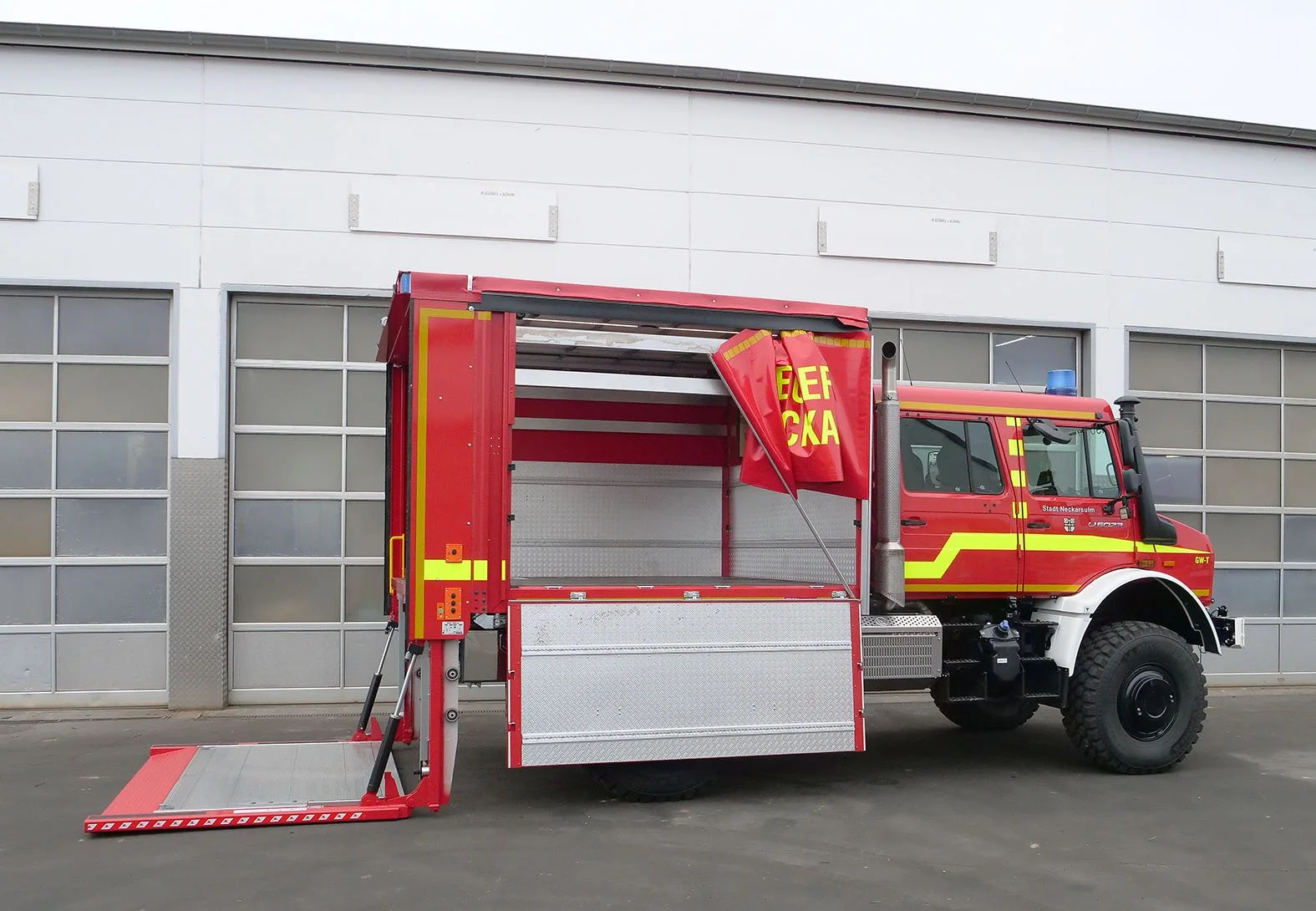 Feuerwehrfahrzeug mit aufklappbarer Rampe, vor einer modernen Gebäude mit großen Fenstern.