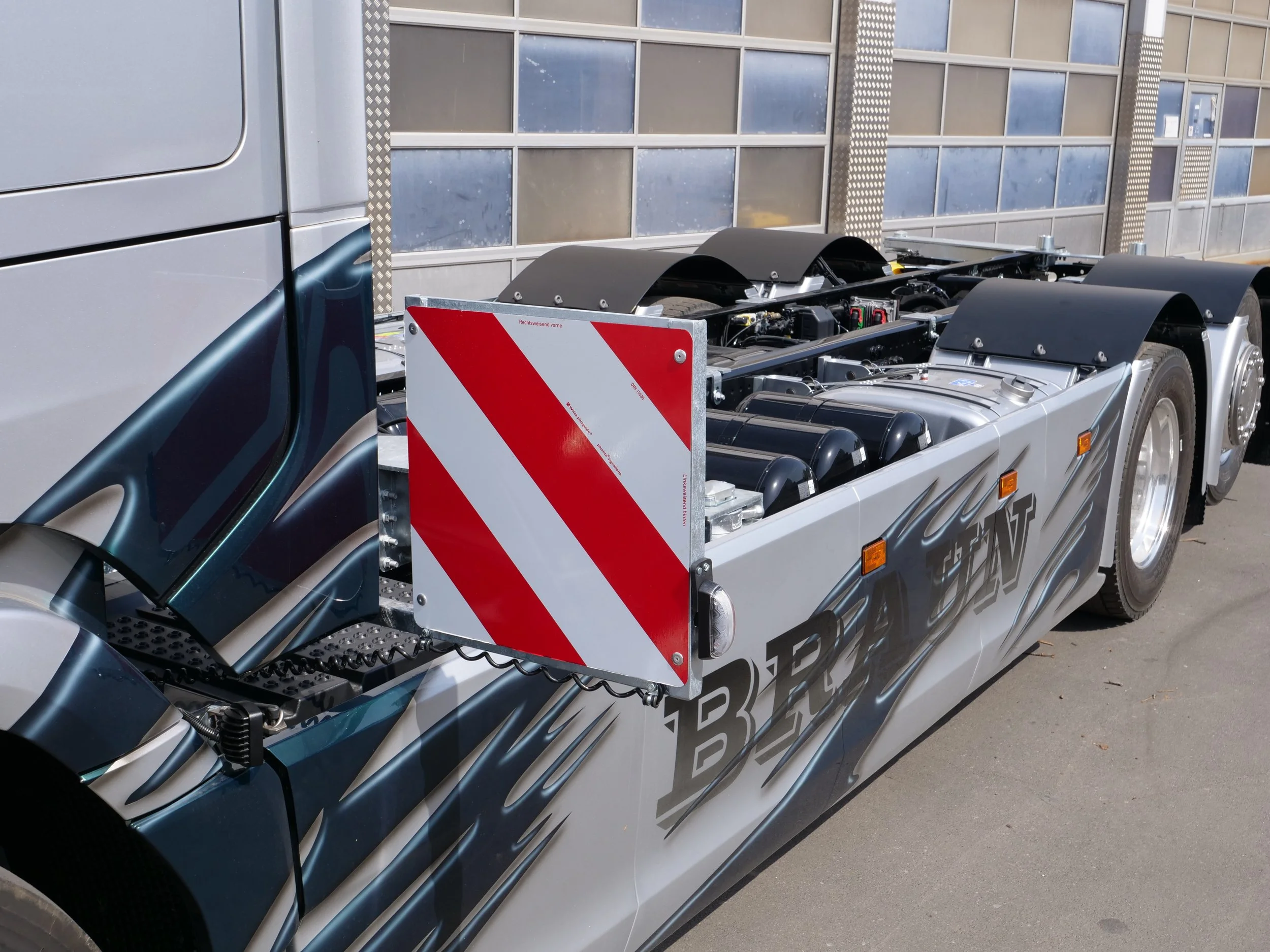 Nahaufnahme eines LKW-Teils, offensichtlich die Antriebsachse, mit schwarzen Reifen und Metallstrukturen, vor einem Gebäude mit großen Fenstern.