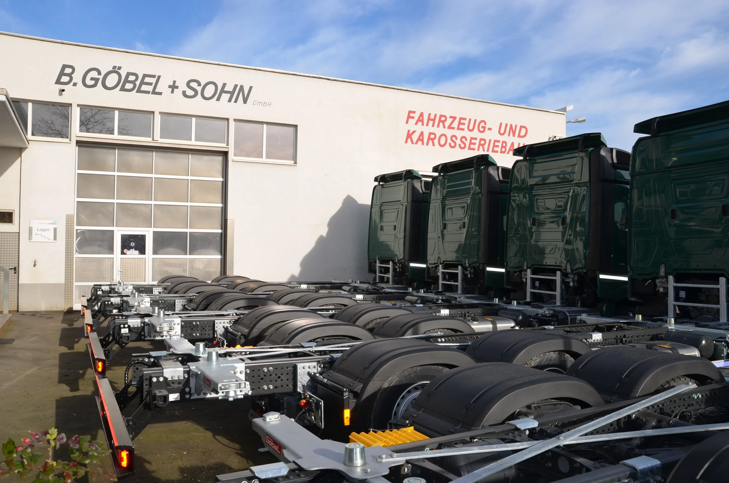 Außenansicht eines Gebäudes mit Firmenname 'B. GÖBEL + SOHN' und großer Garage. Im Vordergrund stehen mehrere leere Fahrzeugrahmen und Radträger. An der Seite steht ein Schild 'Lager'.