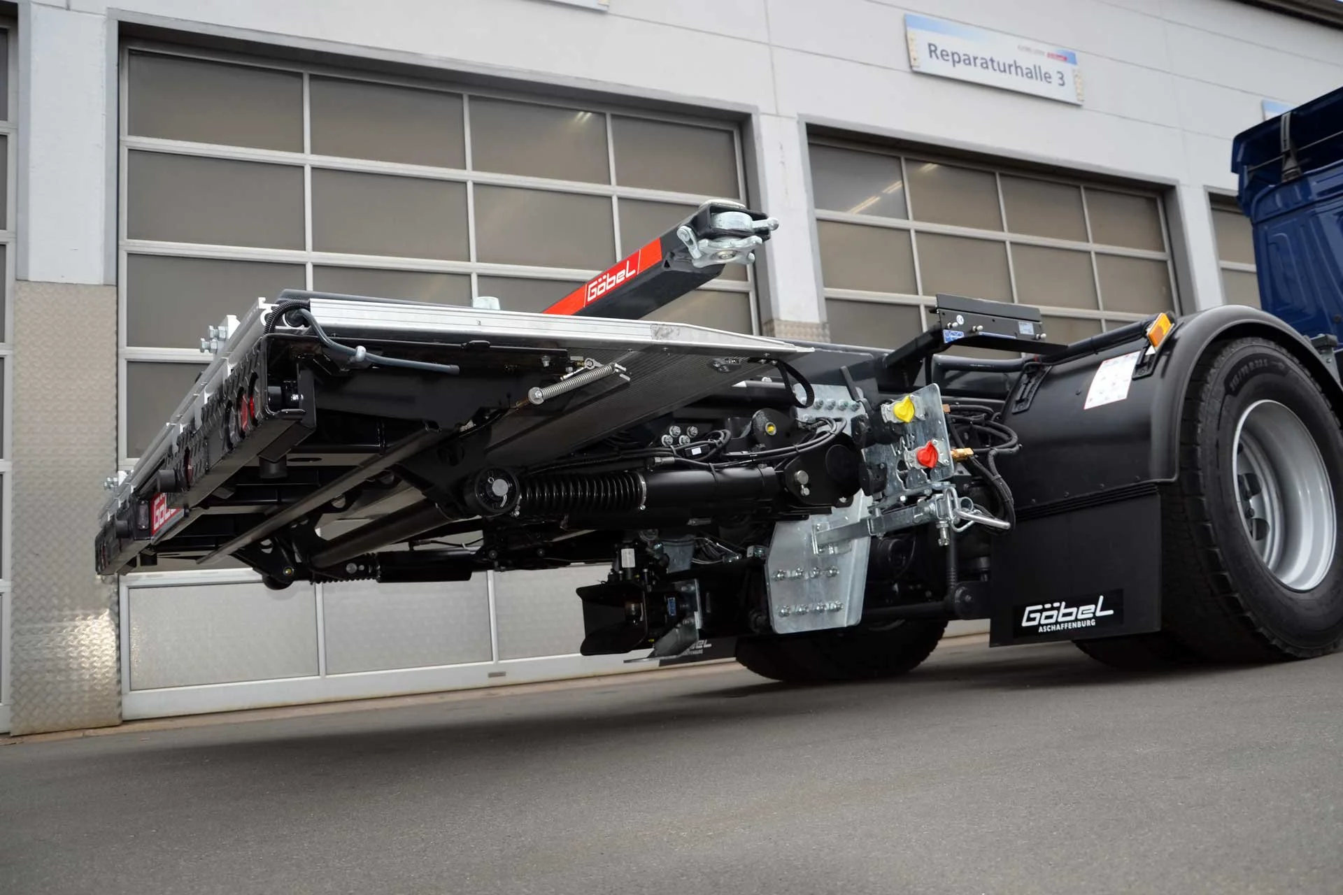 Nahaufnahme eines Fahrzeugchassis mit hydraulischen Komponenten, Reifen und Hydraulikzubehör vor einer Garage, Beschriftung "Gabel" auf den Teilen, Gebäude mit Fenster in Hintergrund.