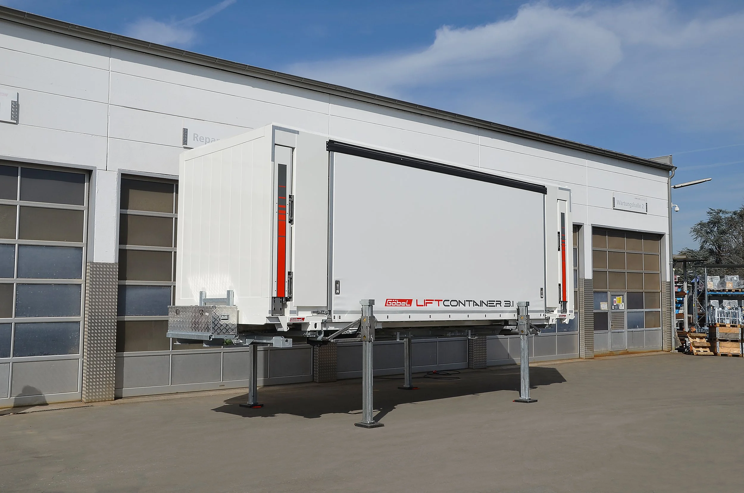 Ein weißer Transportcontainer mit der Aufschrift 'Gabel Lift Container 3.1' ist auf einem mobilen Hebe- und Stützgestell vor einem modernen weißen Gebäude mit großen Fensterflächen abgestellt. Der Himmel ist teilweise bewölkt.