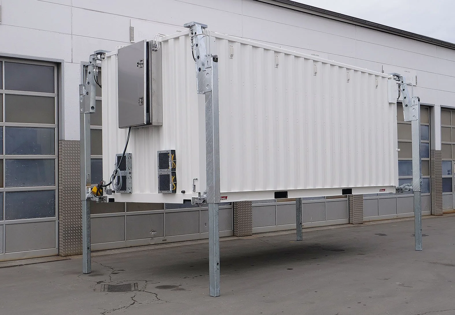 Ein weißer Container, der an einem Gerüst befestigt ist, wahrscheinlich für technische oder industrielle Zwecke, vor einer modernen Gebäude Fassadent mit Fenstern.