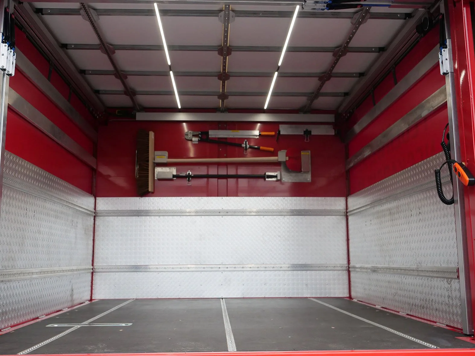 Innensicht eines leeren Feuerwehrautos, mit Werkzeugen an der Wand, darunter ein Besen, eine Schaufel, und ein hydraulisches Rettungsgerät.