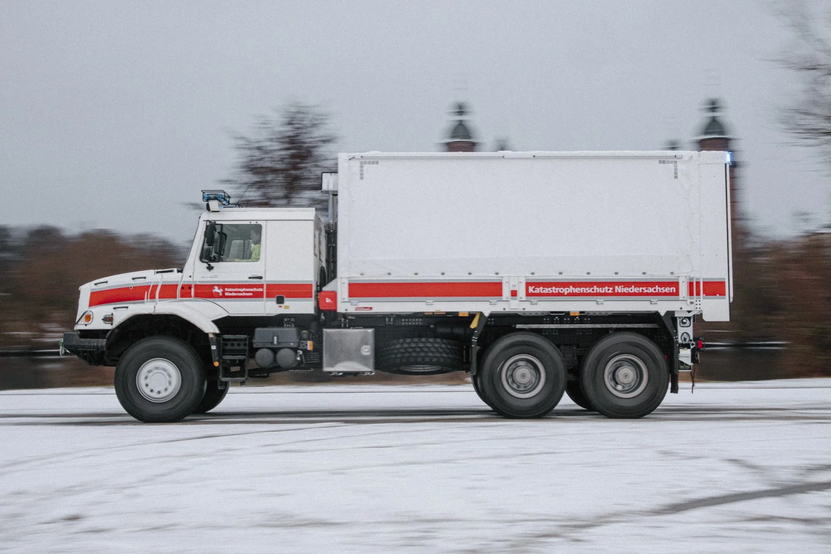 Ein Einsatzfahrzeug des Katastrophenschutzes Niedersachsen fährt auf einer schneebedeckten Straße, im Hintergrund sind Bäume und eine Kirche zu sehen.