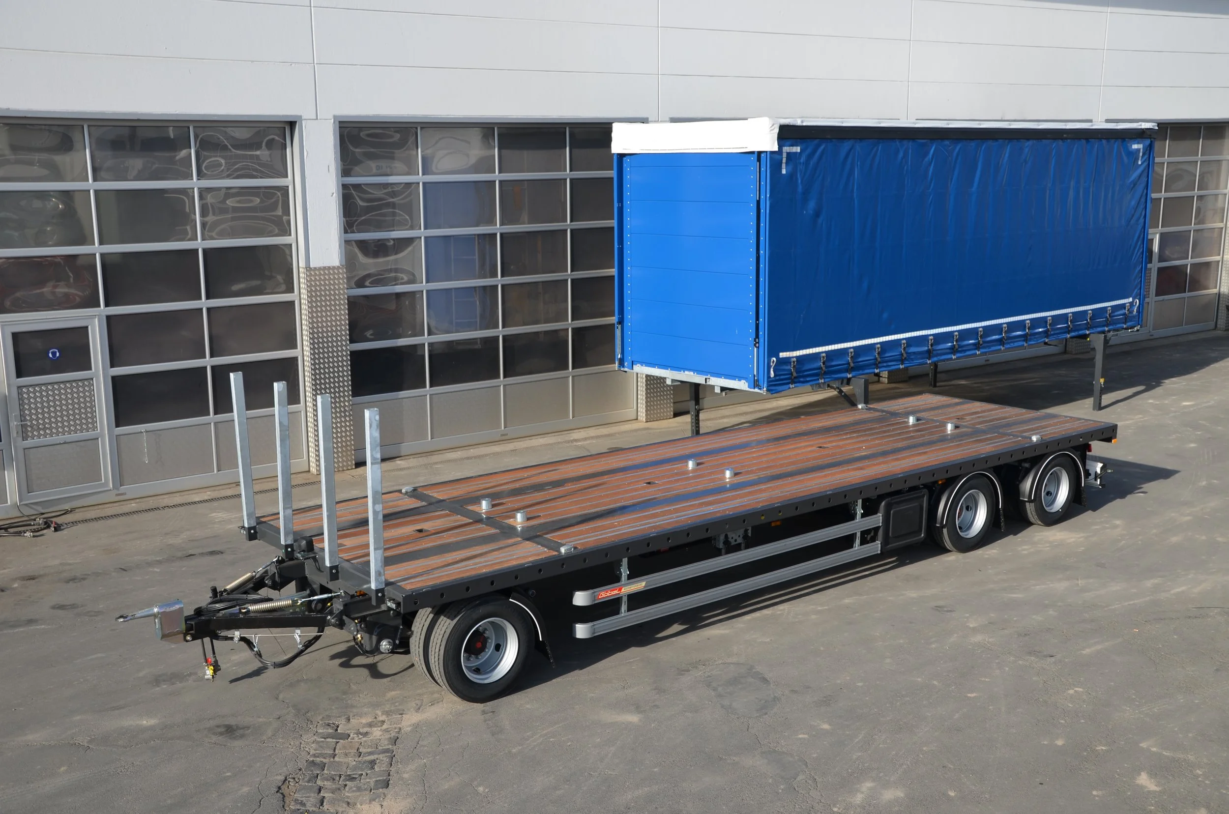 Ein blauer Transportwagen mitseiten, auf einem Anhänger, vor einem modernen Gebäude mit großen Fensterfronten.