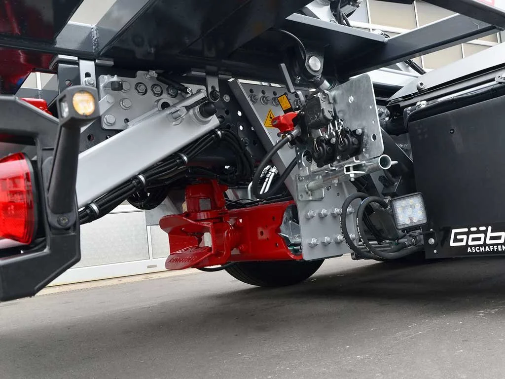 Nahaufnahme der Unterseite eines Fahrzeugs mit technischen Komponenten, Hydraulikleitungen, elektronischen Teilen und Beschriftungen in Deutsch.