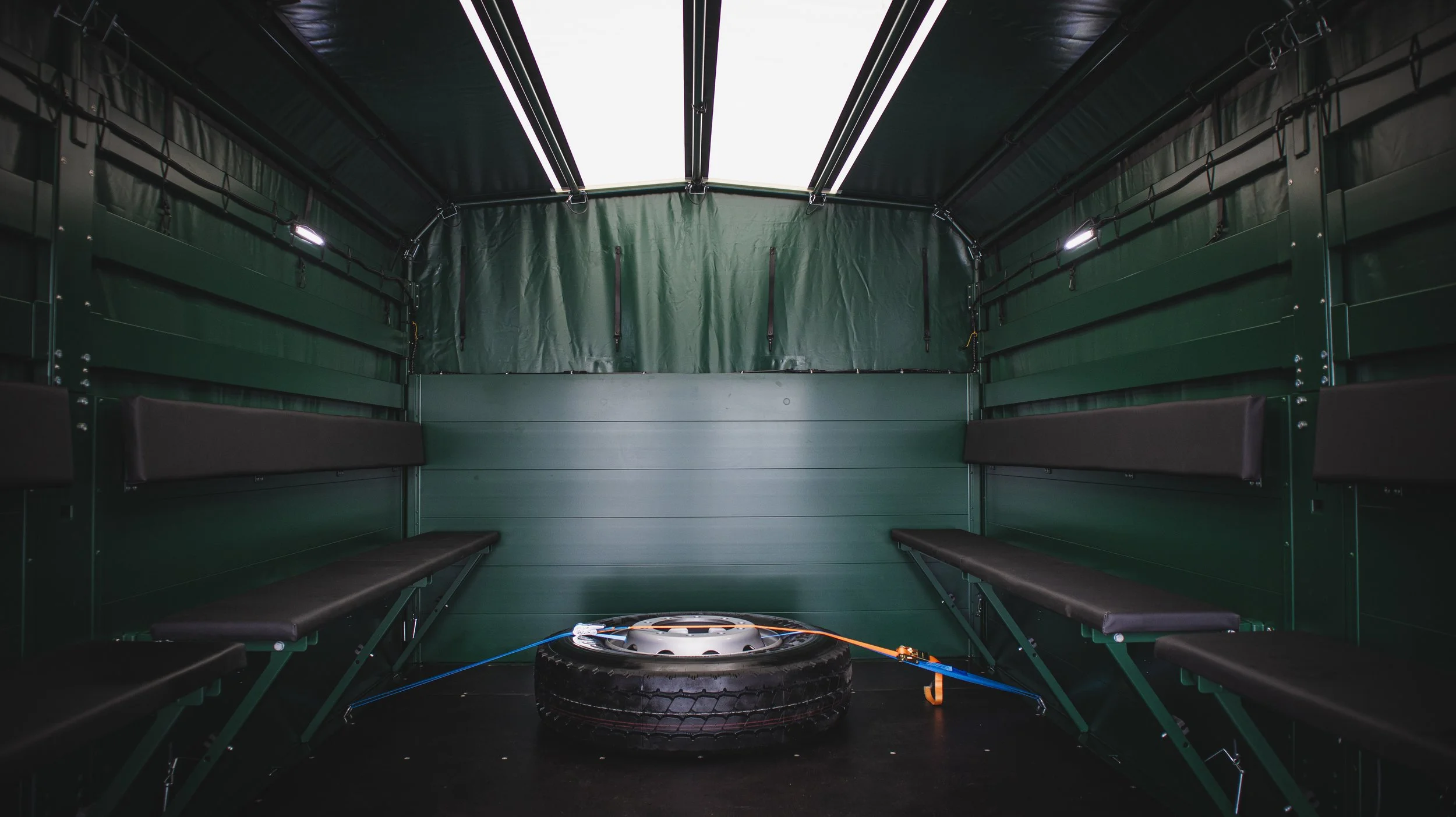 Innenansicht eines grünen IVECO Eurocargos LKW als Truppentransportfahrzeuges. Mit schwarzen Polsterbänken an den Seiten, einem Reifen in der Mitte und elektrischen Anschlüssen. Aufgebaut durch die Firma Göbel Fahrzeugbau.